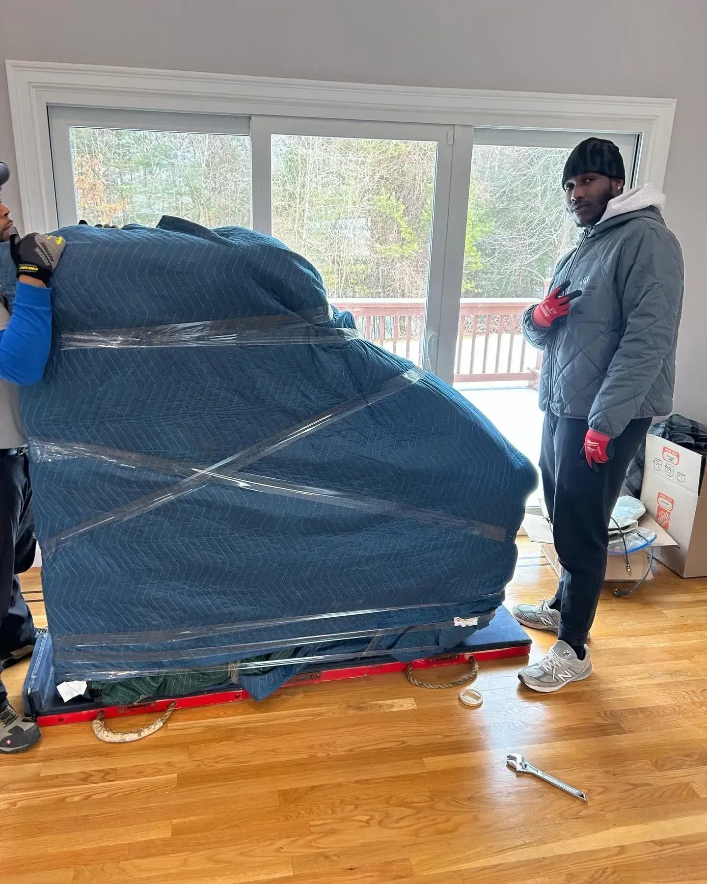 Piano being moved inside. Men wrap it with blue blankets and tape near a sliding glass door.