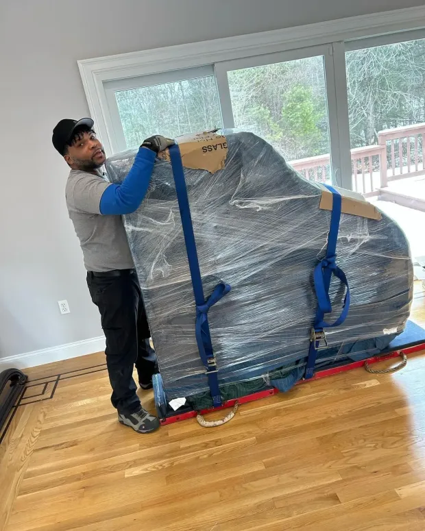 Man moving a wrapped piano on a dolly with blue straps in a room with hardwood floors and a window.