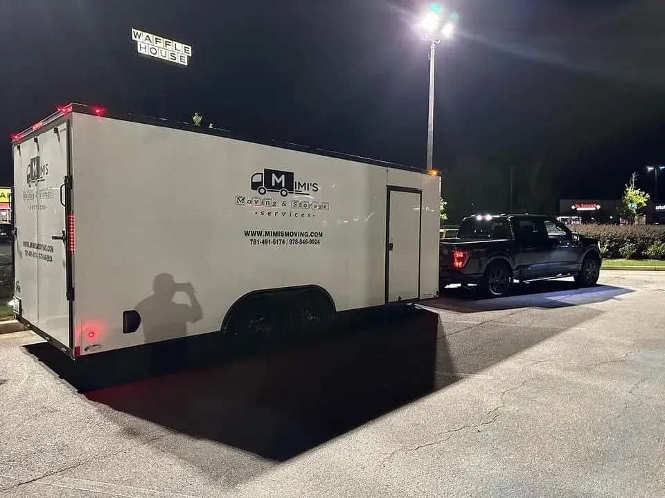 A white trailer with black lettering is attached to a black truck in a parking lot at night.