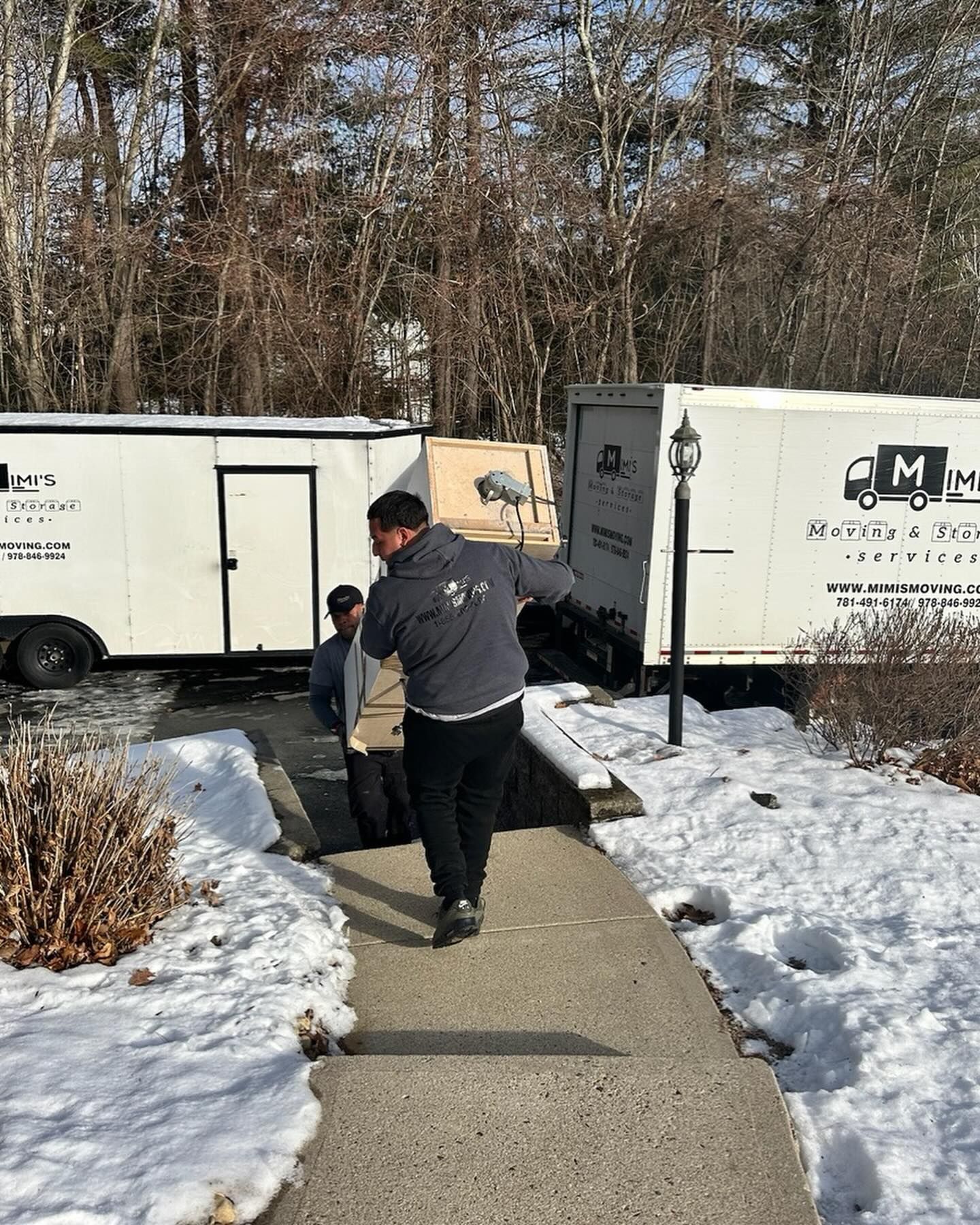 Two movers carrying a large box toward a truck on a snowy walkway. A second truck and trailer are visible.