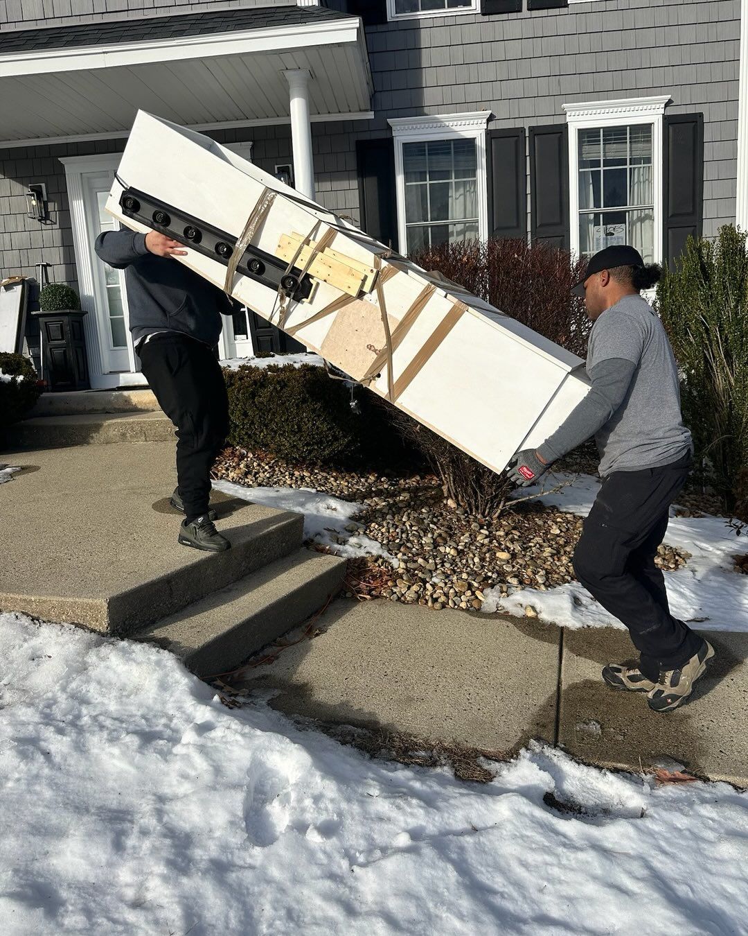 Two people carrying a large, white object up steps in front of a house on a snowy day.