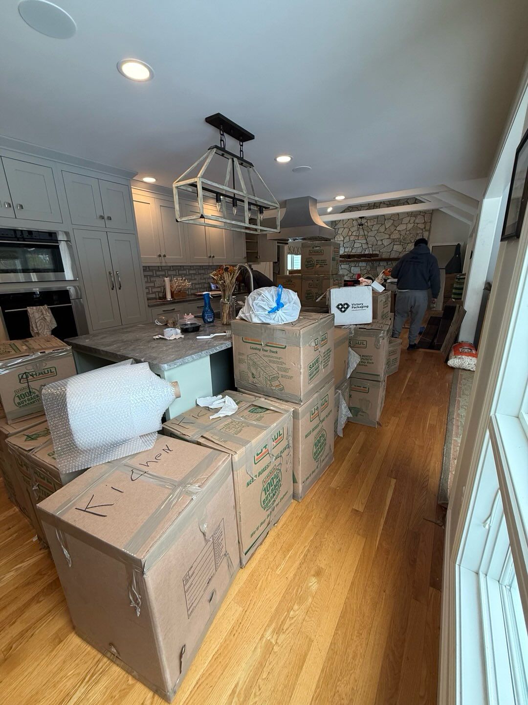 Boxes packed for a move in a kitchen with a person in the background. Wooden floors and white cabinets.