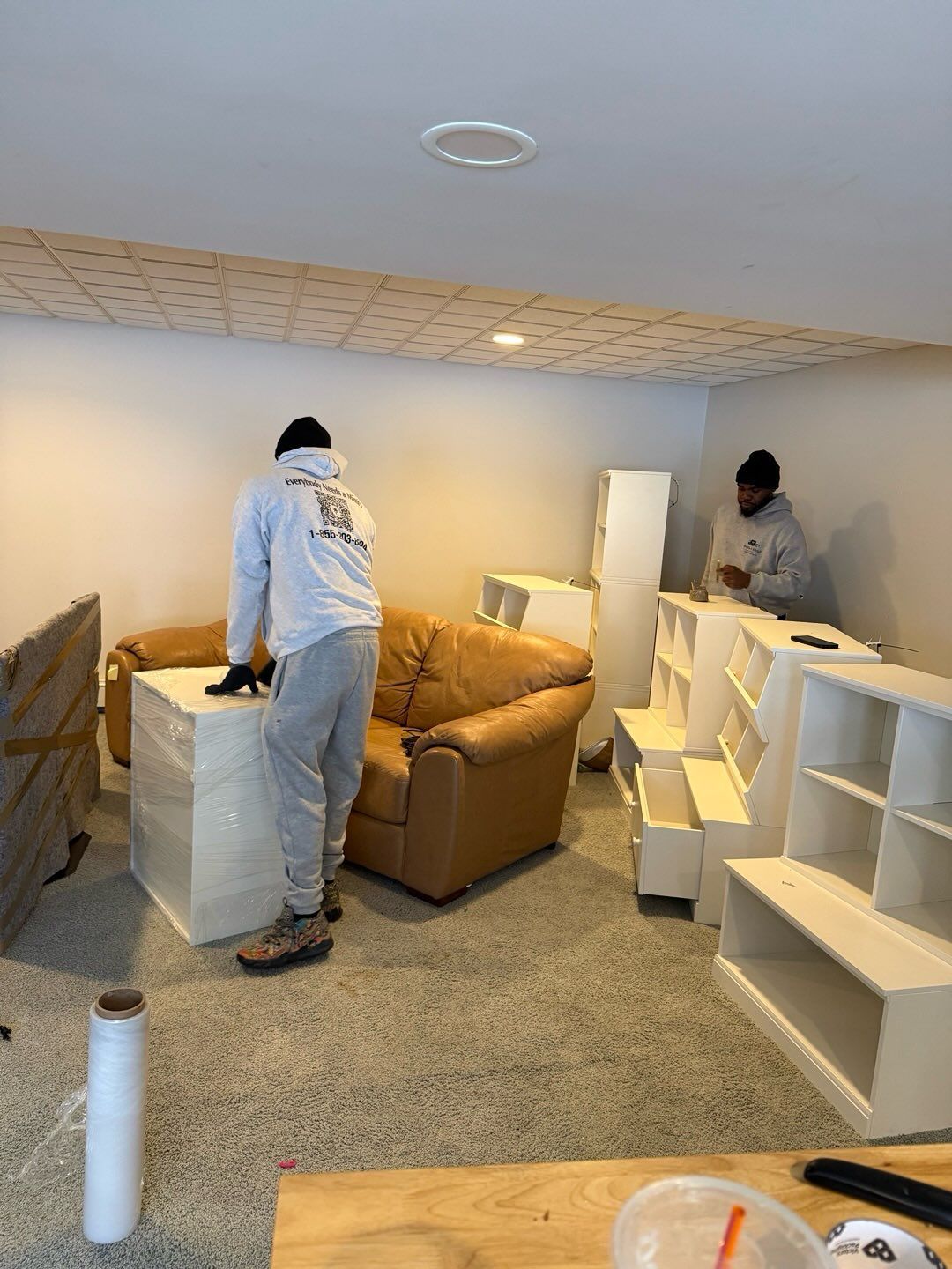 Two people wrapping furniture in a basement room. White shelves and a tan chair are present.