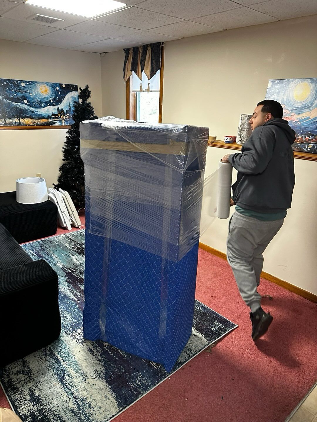 Person carrying roll of packing material next to a large, wrapped blue object on a rug. Interior setting.