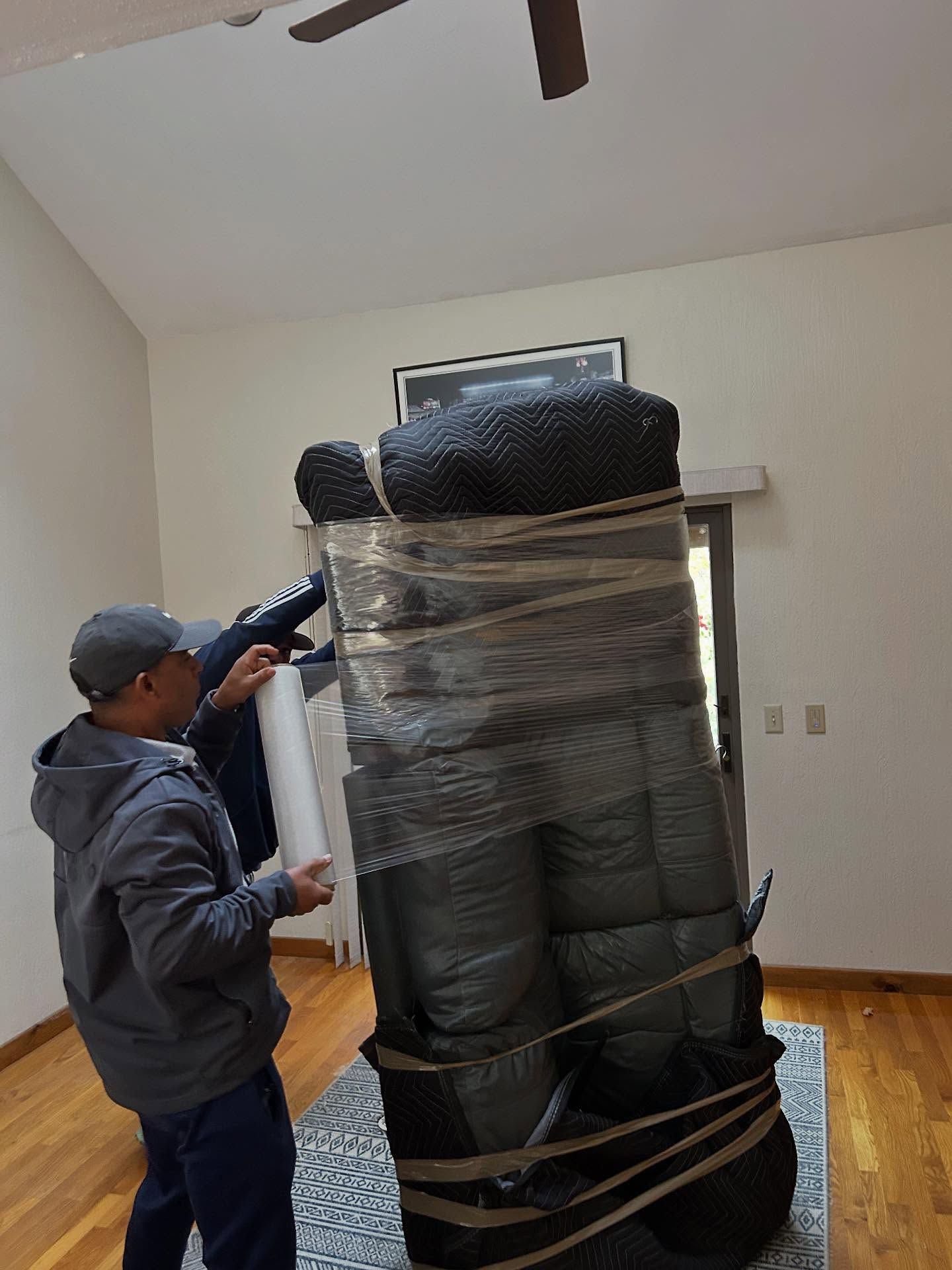 Two people wrapping a large, black couch with plastic wrap in a room with hardwood floors and a rug.