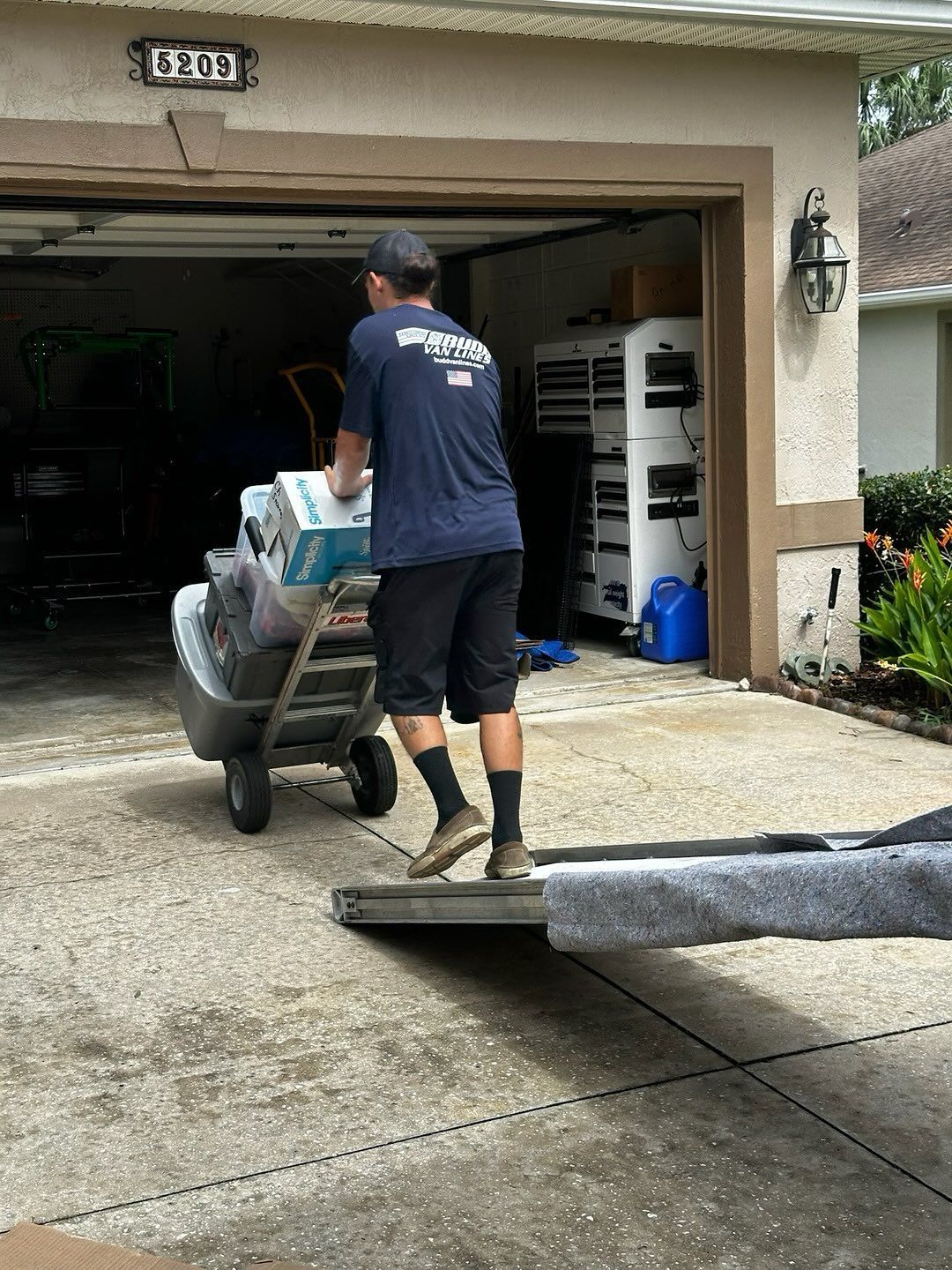 Man pushing a dolly loaded with boxes out of a garage onto a driveway.