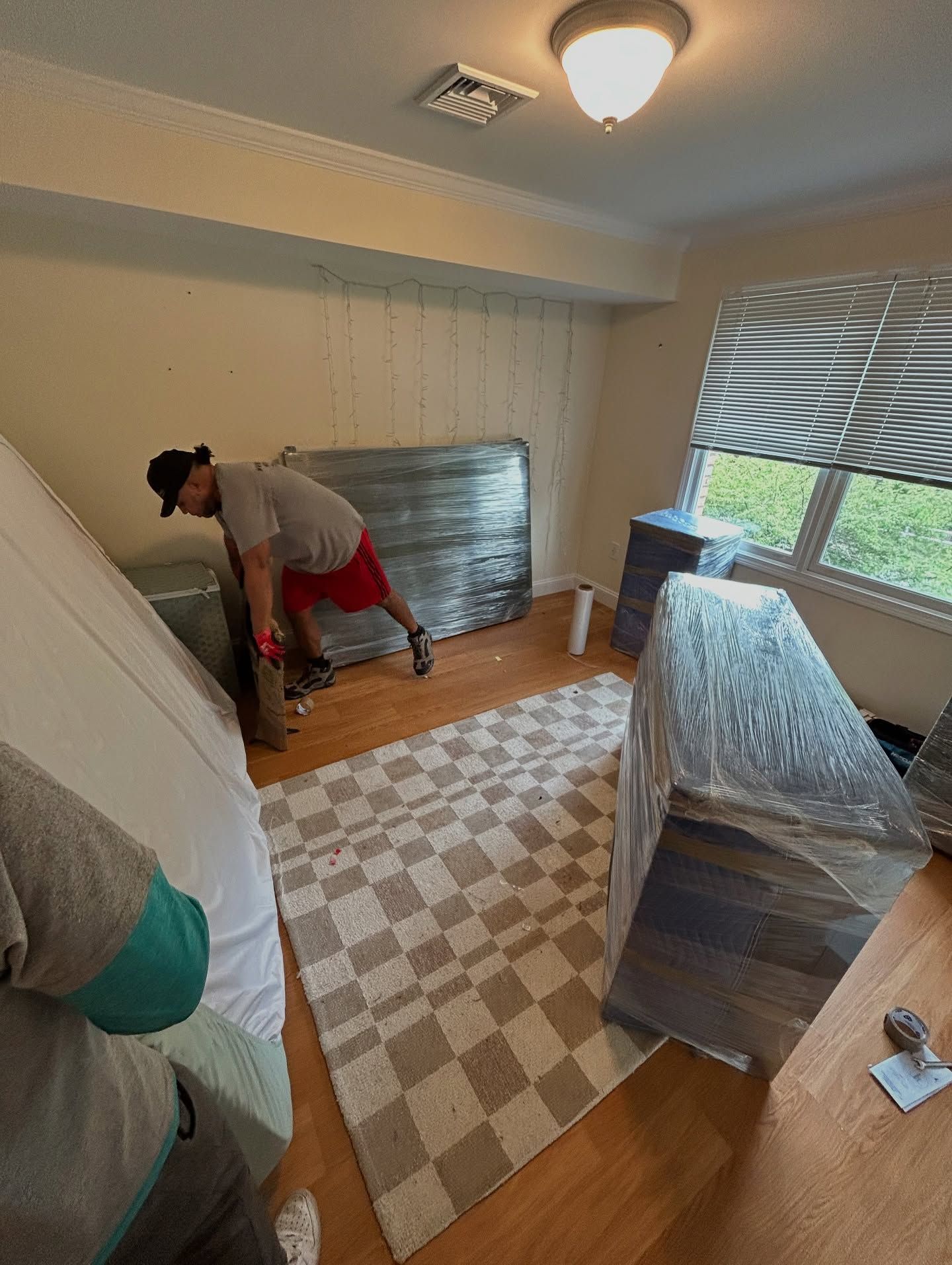 Person in red shorts moving furniture in a room. Boxes and a mattress are wrapped. Brown and white rug on wood floor.