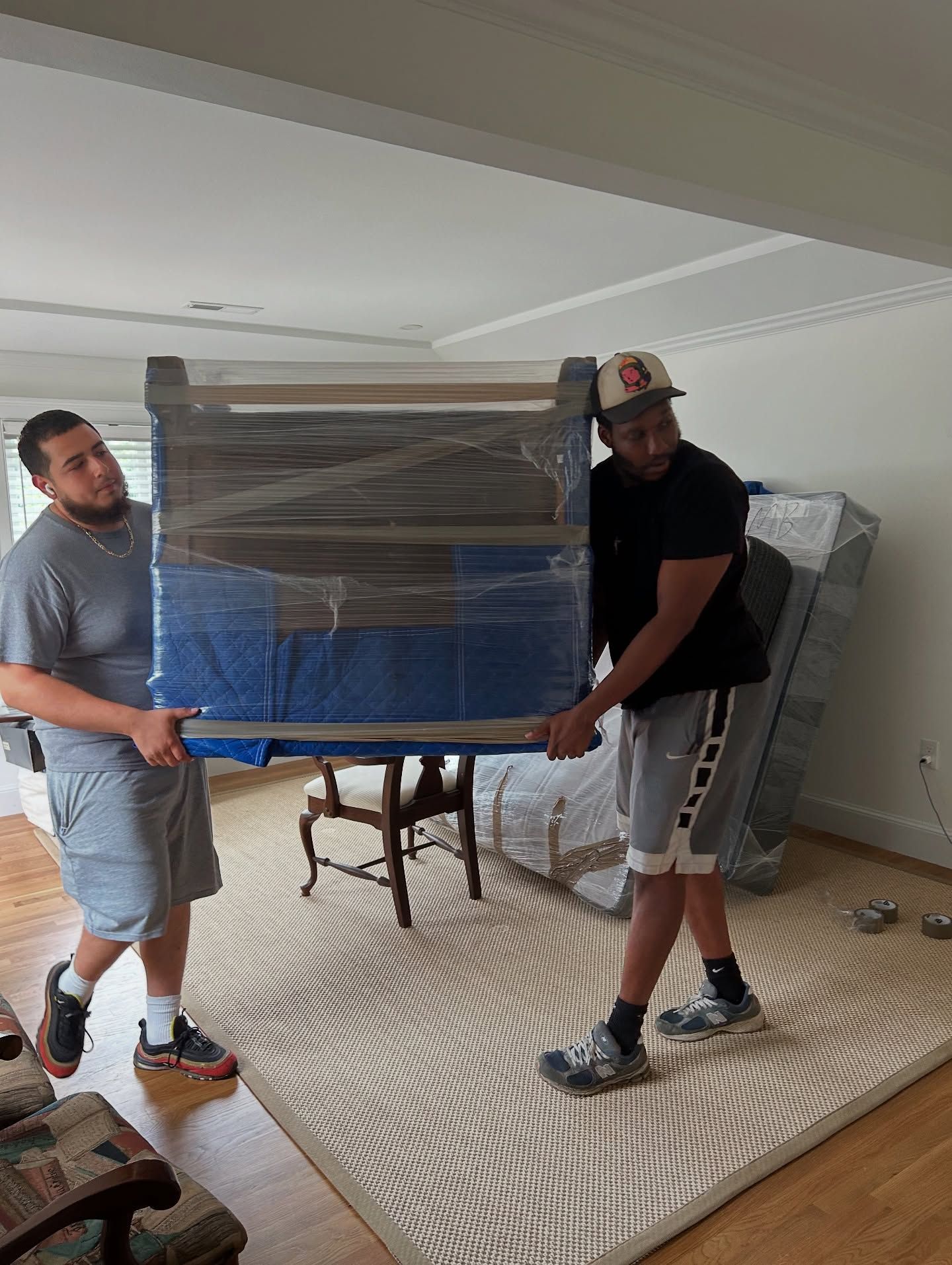 Two people carrying wrapped furniture in a room with hardwood floors and a rug.
