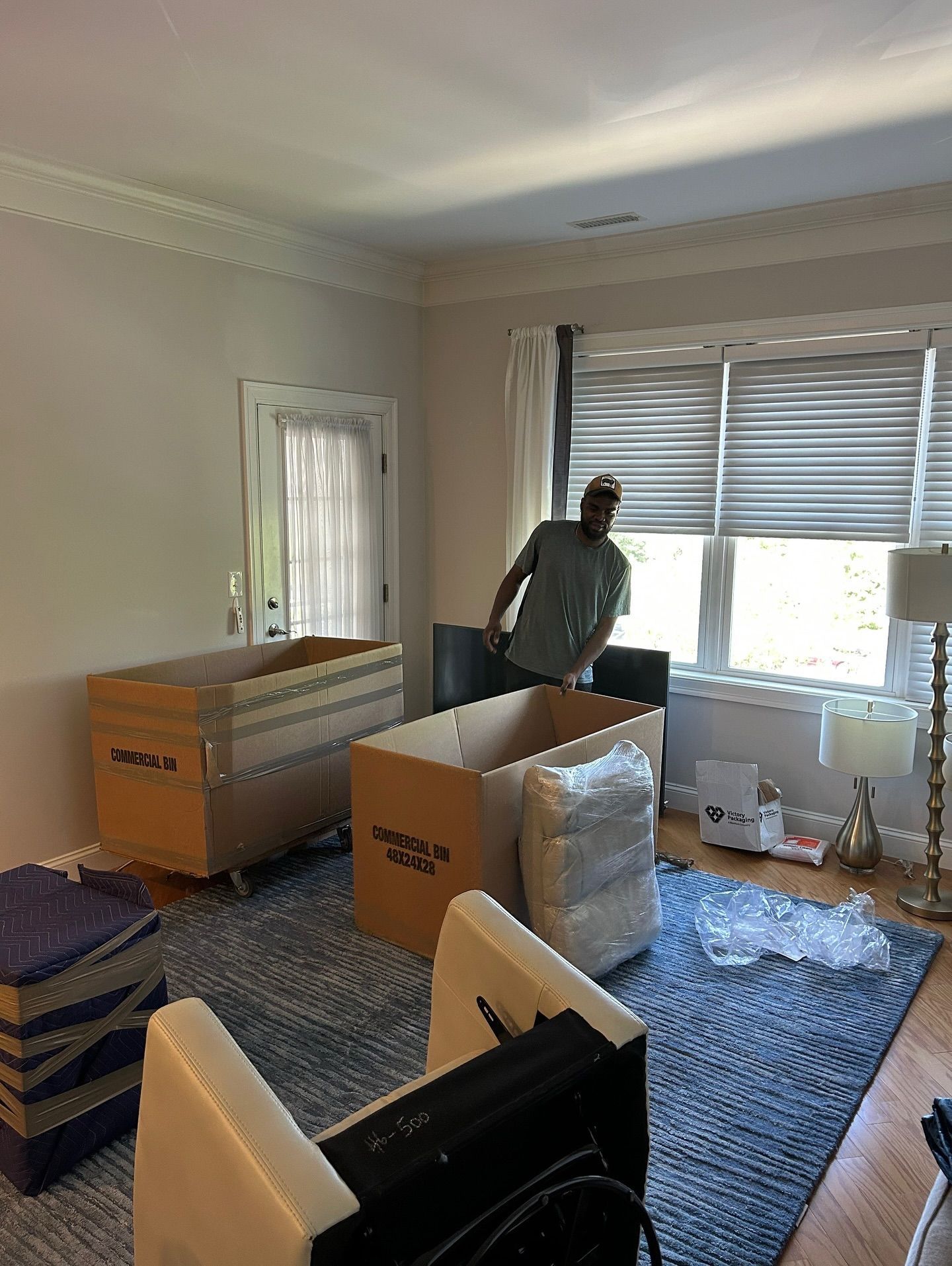 Person in a room with moving boxes, looking toward a window.  Boxes, furniture, and a rug are visible.