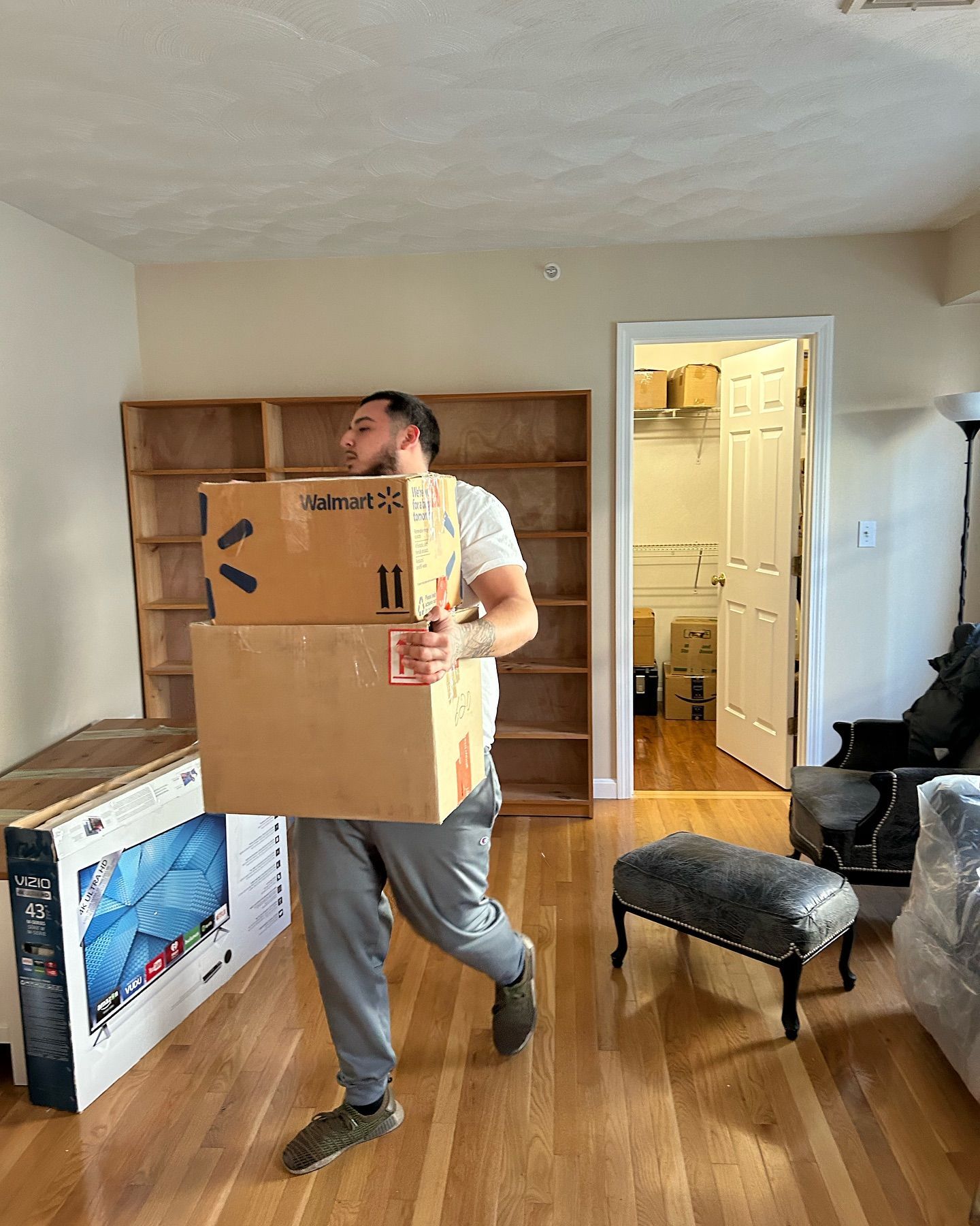 Man carrying boxes in a room with wood floors. Boxes and a TV box are present. A bookcase is against the wall.
