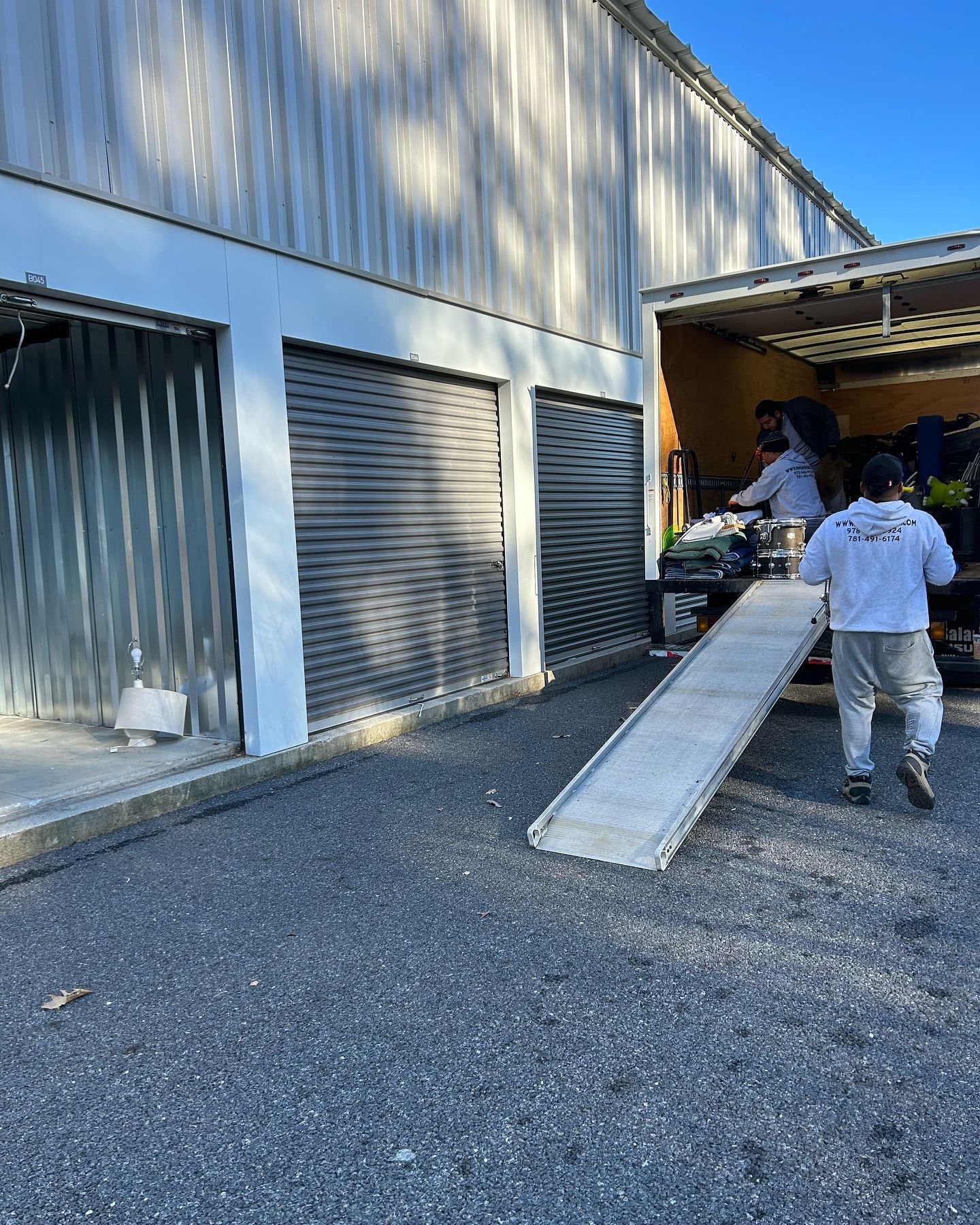 Two people loading a truck from storage units with roll-up doors, using a ramp. Outdoor setting.