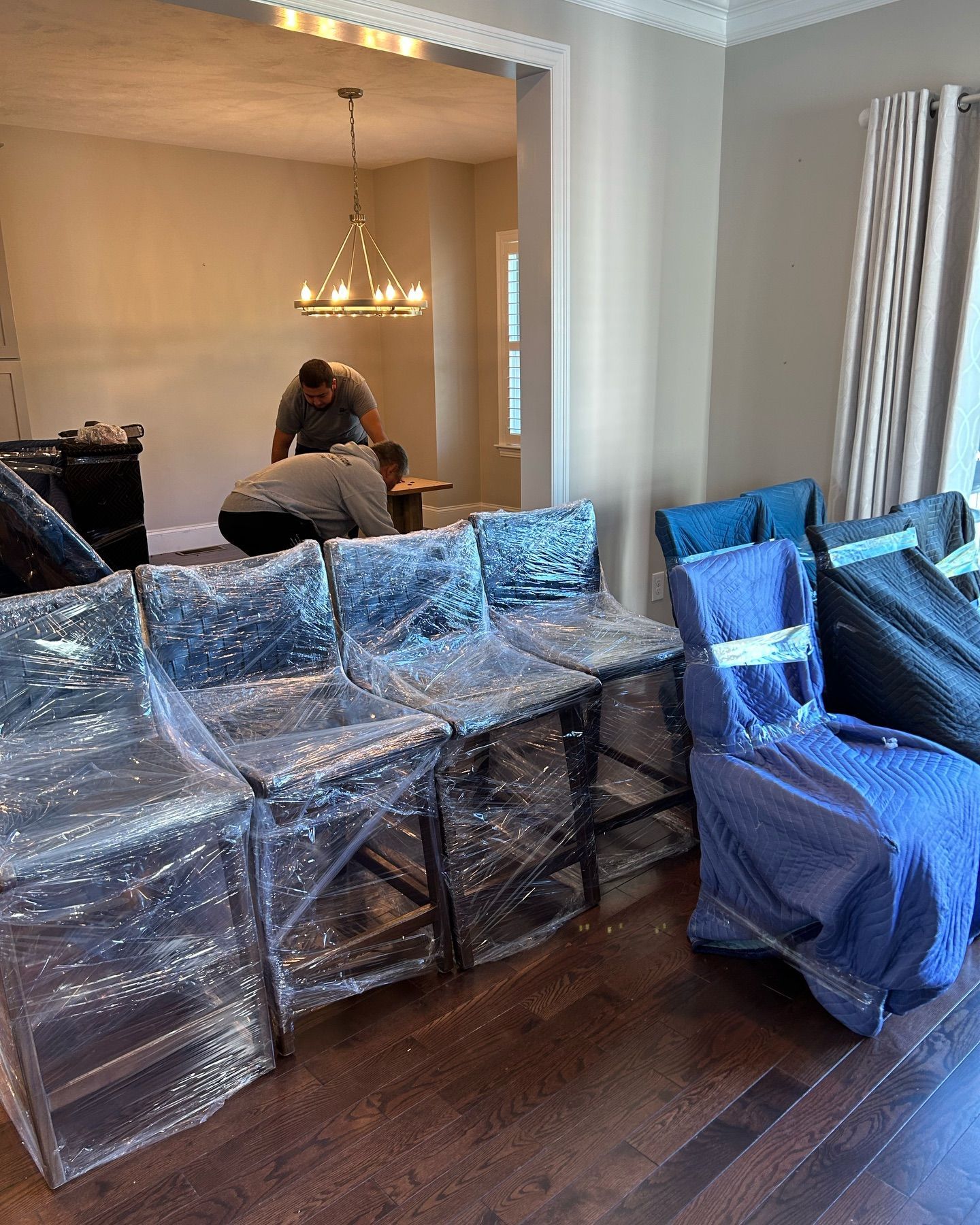 Chairs wrapped in plastic and blue covers; movers working in a room with a chandelier.