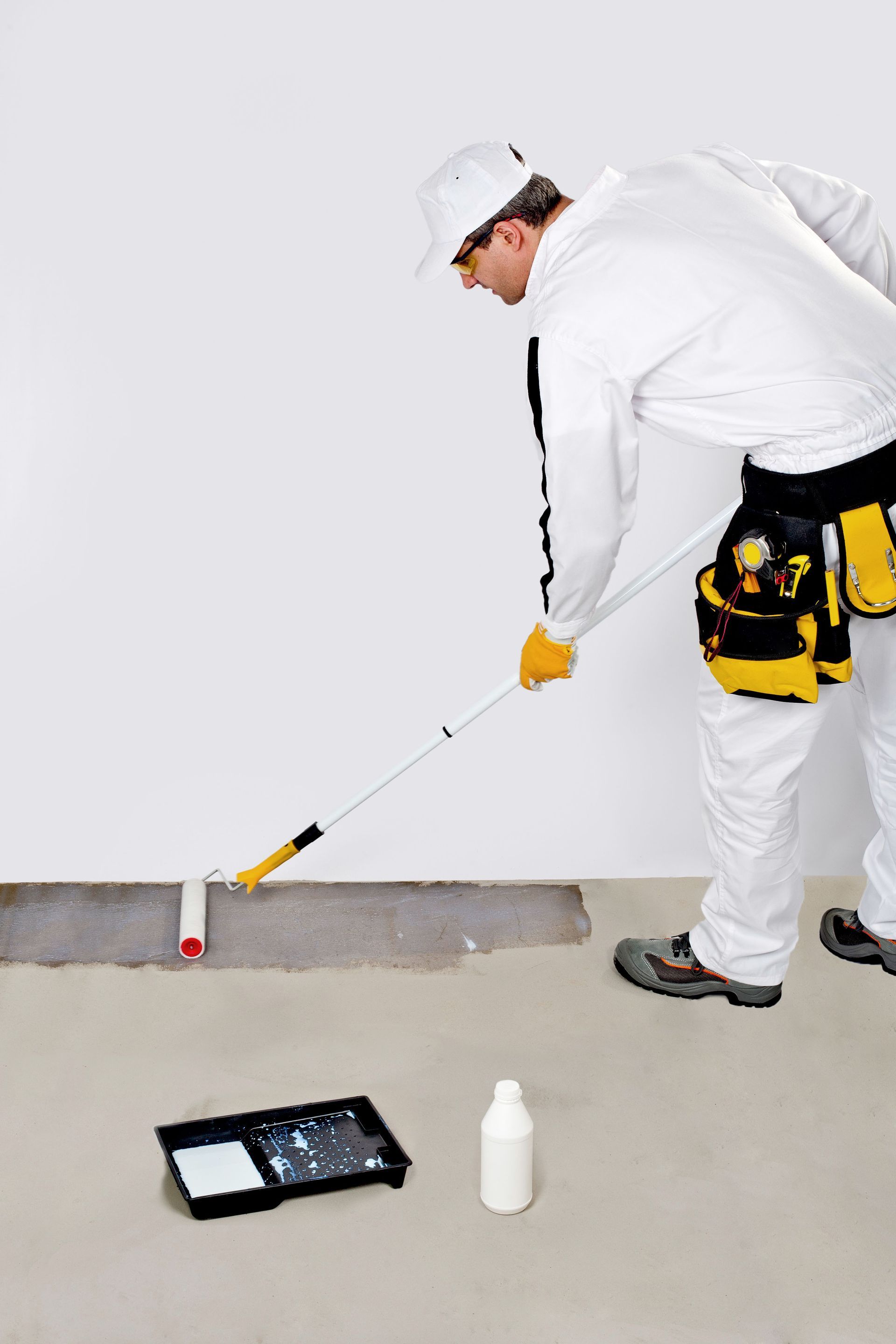 Painter using a roller to apply paint to a wall. He wears white protective clothing and a tool belt.