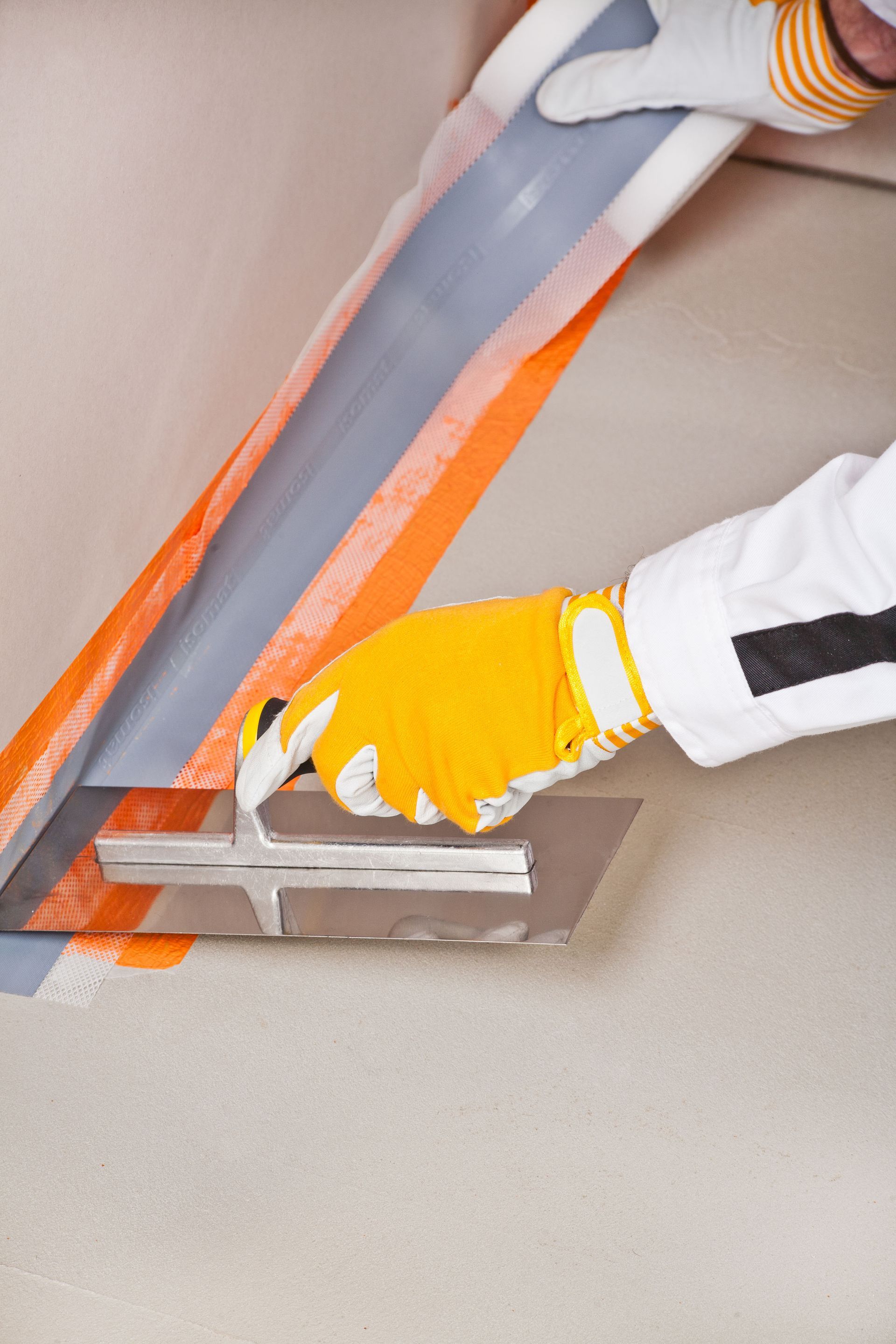 Person wearing gloves smoothing sealant with a trowel, applying orange waterproof tape to a wall.