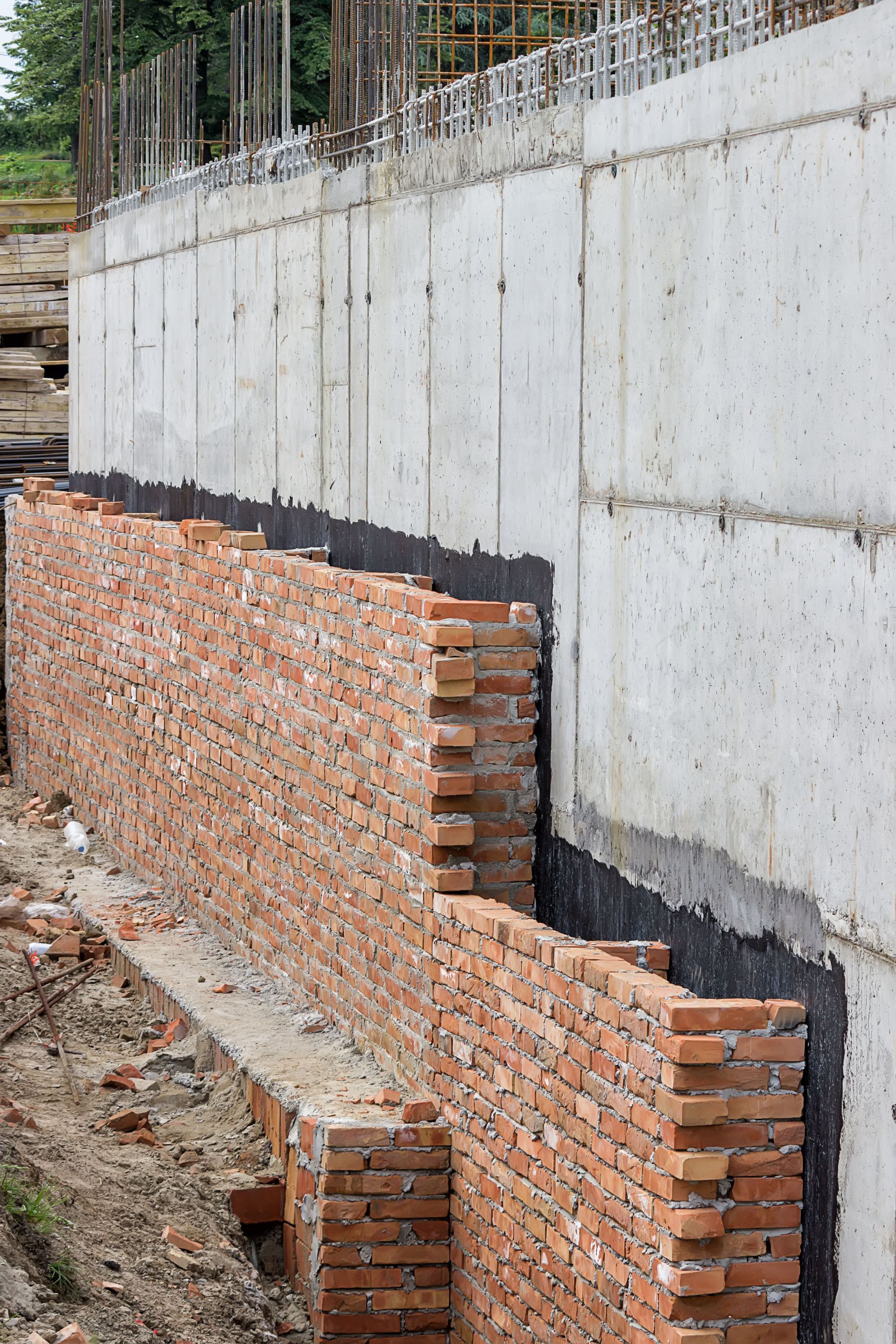 Brick wall being built against a concrete wall at a construction site. Black sealant visible.