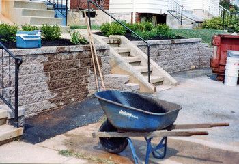 Blue wheelbarrow in front of stone retaining wall and steps leading to a house.
