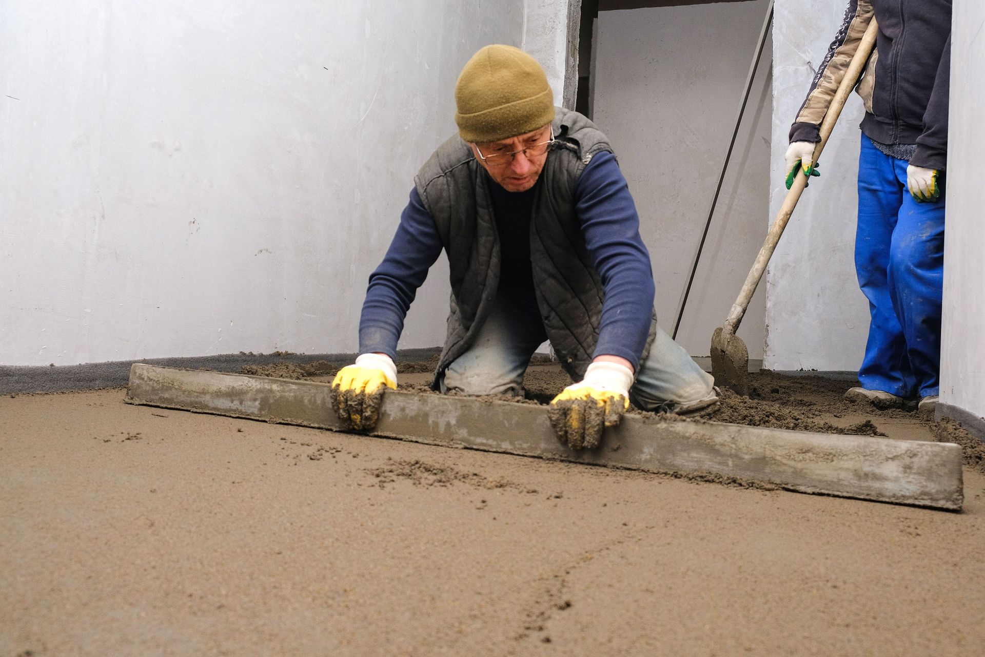 Man leveling concrete floor with a screed; another person holds a shovel.