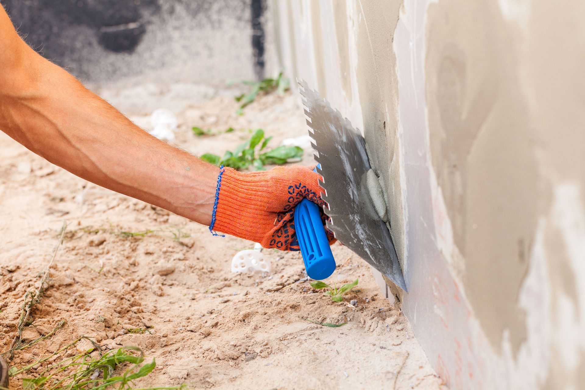 A person using a trowel to apply plaster to a wall outdoors, wearing orange work gloves.
