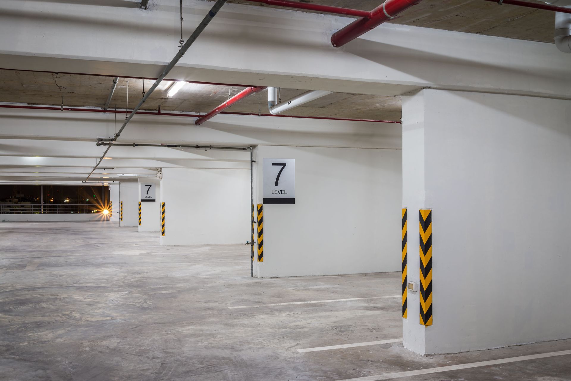 Empty parking garage with white pillars, overhead pipes, and parking space markers.