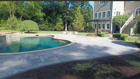 Swimming pool with paved patio, large house, and trees in background.