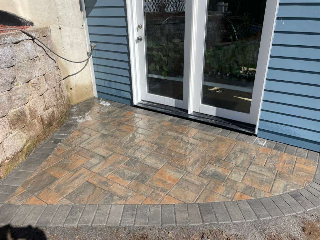 Paver patio in front of blue house with double glass doors, next to a stone wall.