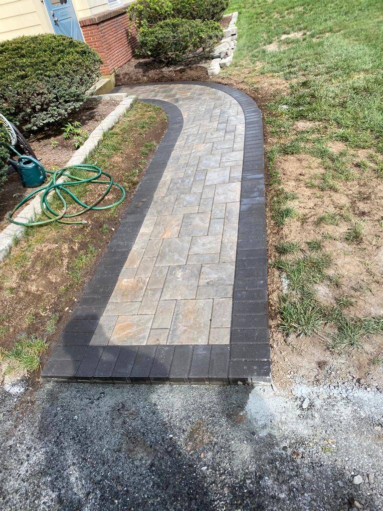 Brick paver walkway with dark border and curved path, leading towards bushes and grass.