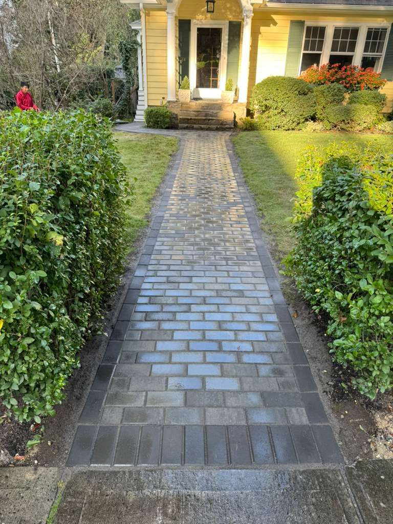 Brick pathway leading to a yellow house with bushes on either side. A person in red is visible on the left.