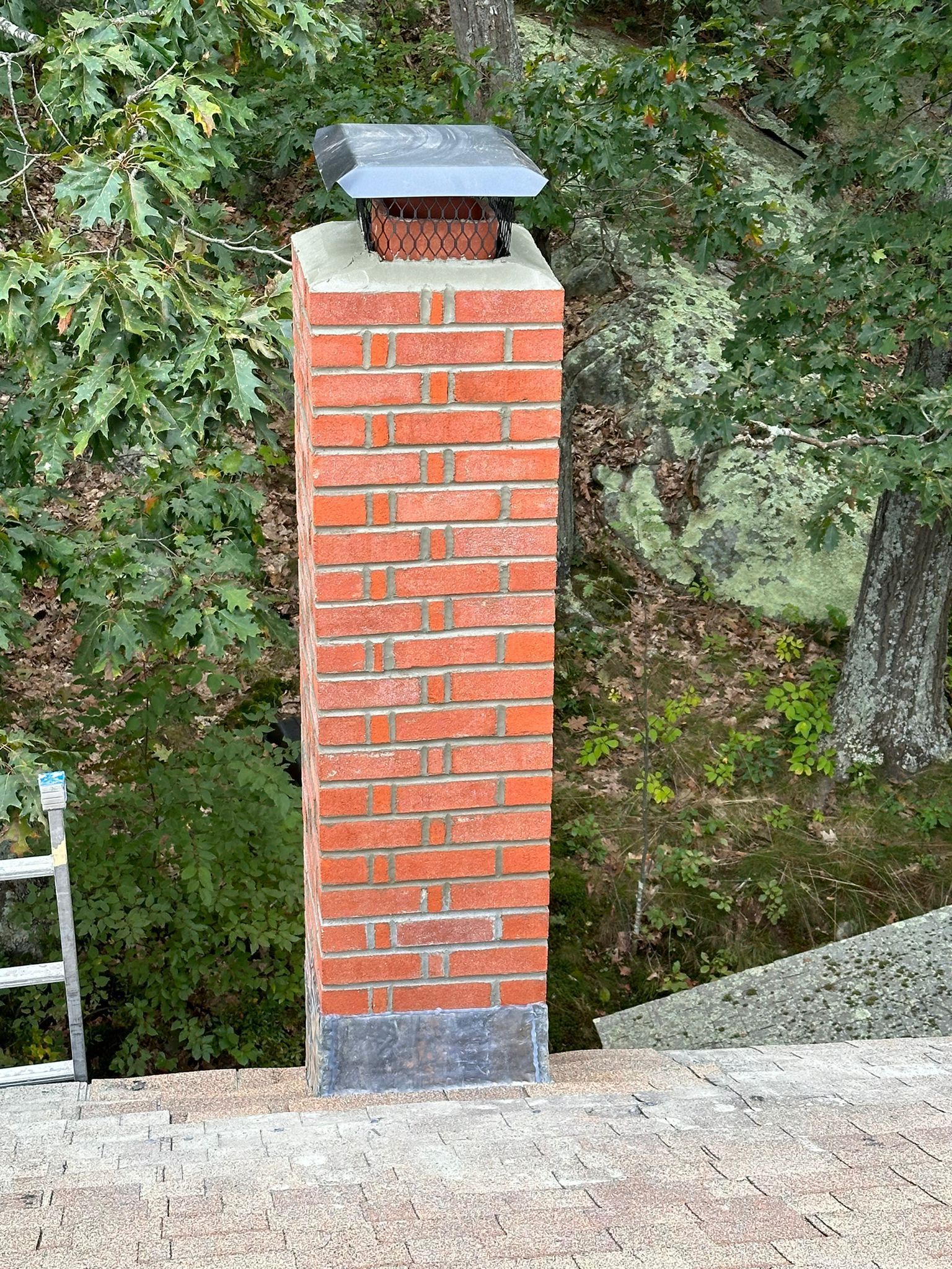 Brick chimney with metal cap, on a roof with trees in the background.