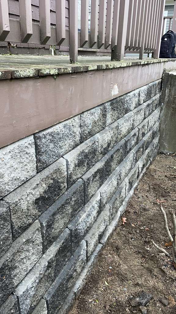 Stone retaining wall supporting a deck. Gray and black blocks, brown deck with railing.