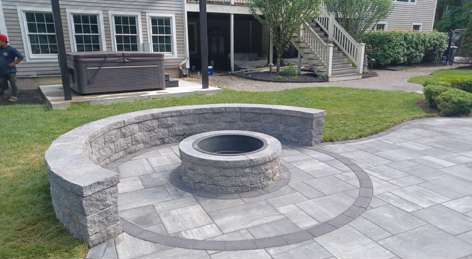 Circular stone patio with fire pit, curved stone wall, and lawn in a backyard.