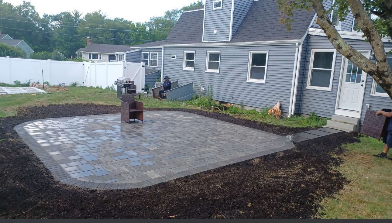 Backyard patio with brick pavers, surrounded by mulch, next to a blue house.
