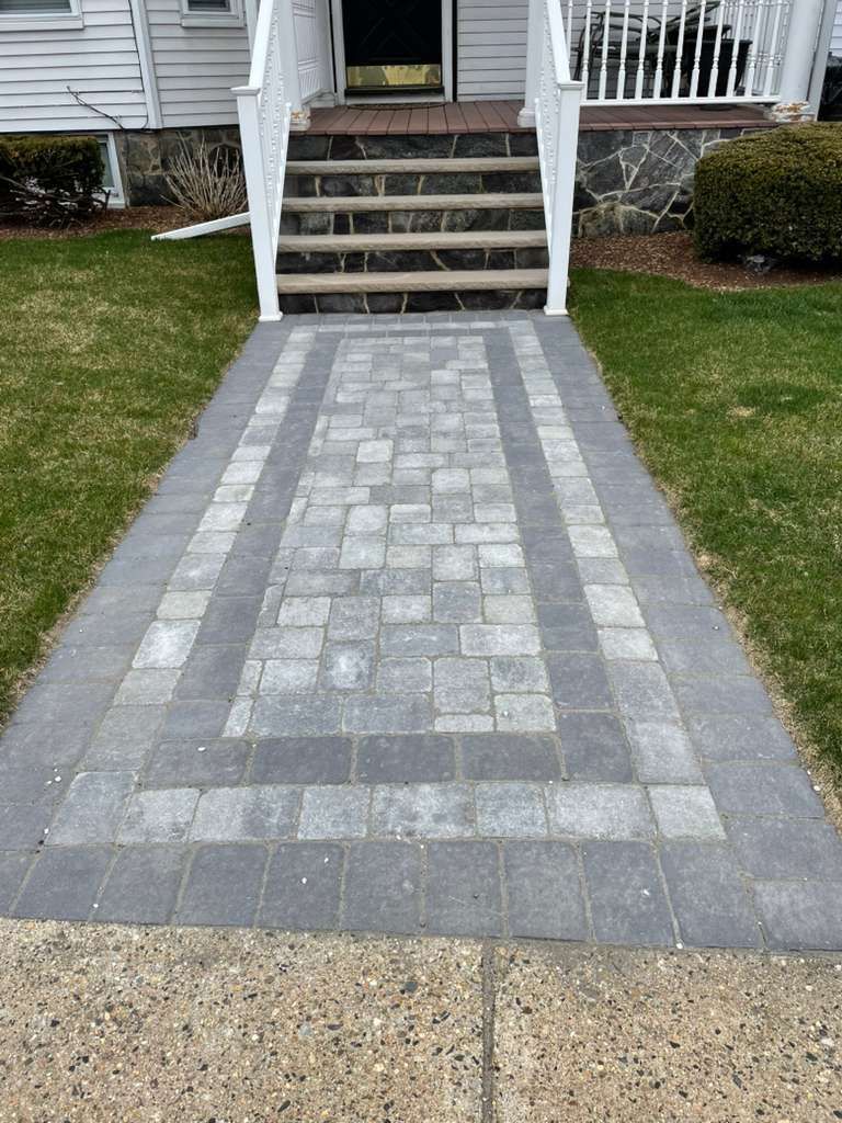 Paver walkway leading to a house entrance with stairs. Dark gray and light gray bricks.