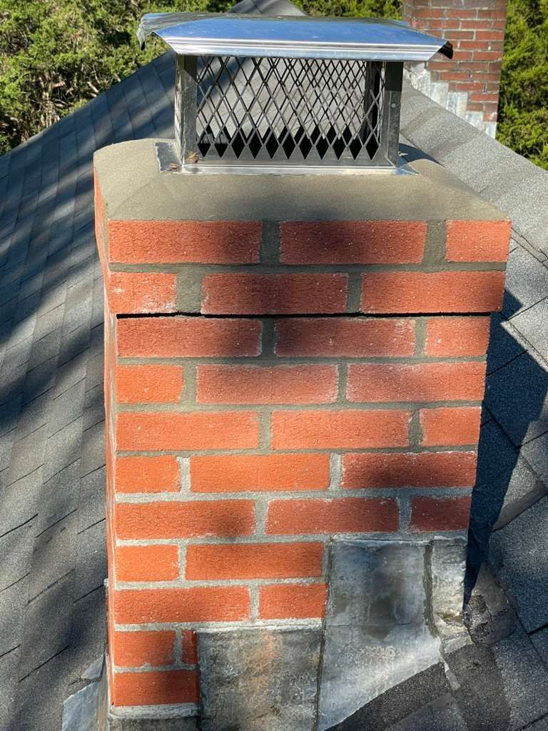 Brick chimney with a metal cap on a rooftop, showing a horizontal crack.