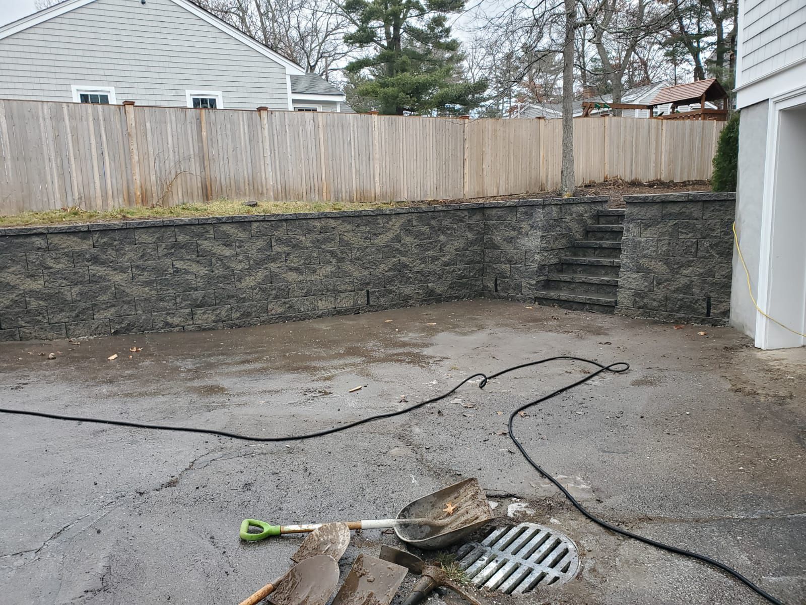 Asphalt driveway with a retaining wall, a water hose, and a drain. A shovel is near the drain.