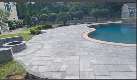 Poolside patio with gray pavers, fire pit, and pool surrounded by grass and trees.