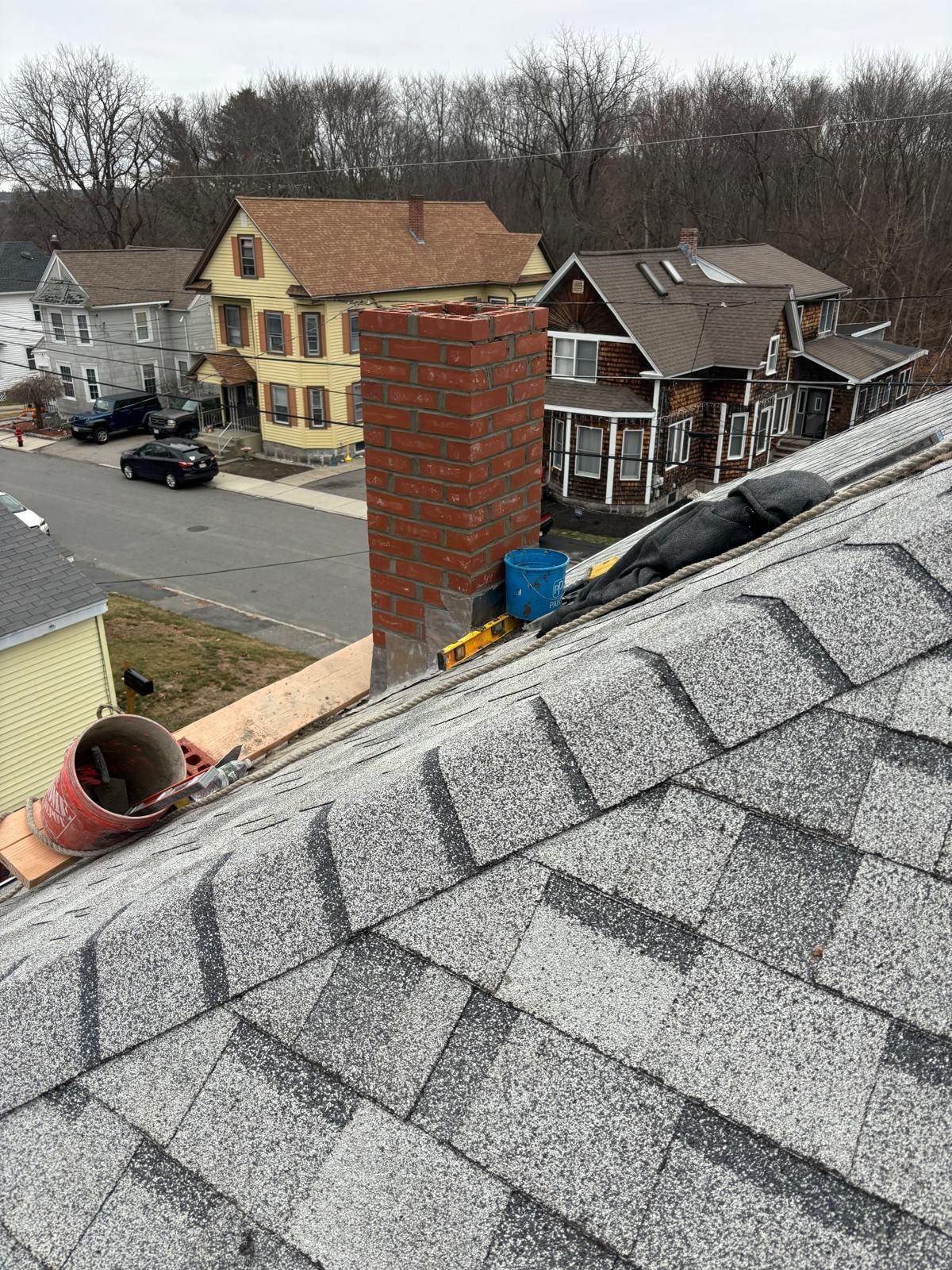 Rooftop view of gray shingles, brick chimney, and houses with cars on a street.