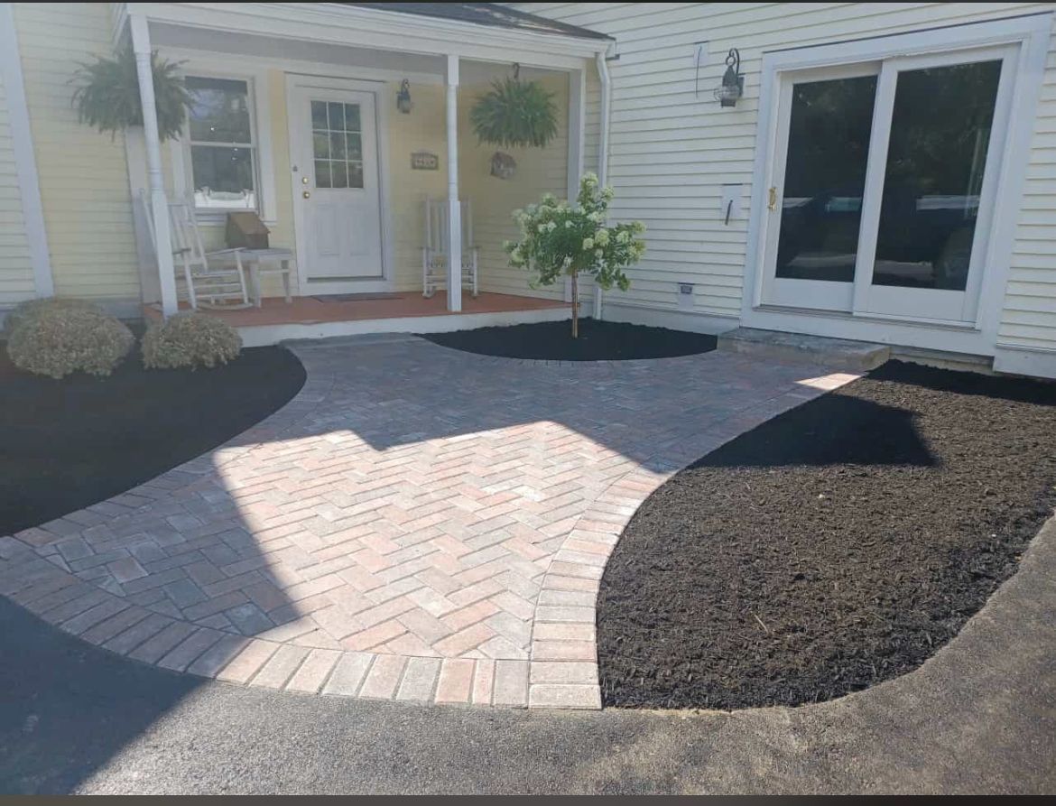 Paver pathway leading to a yellow house with a white door and sliding glass doors, surrounded by black mulch.