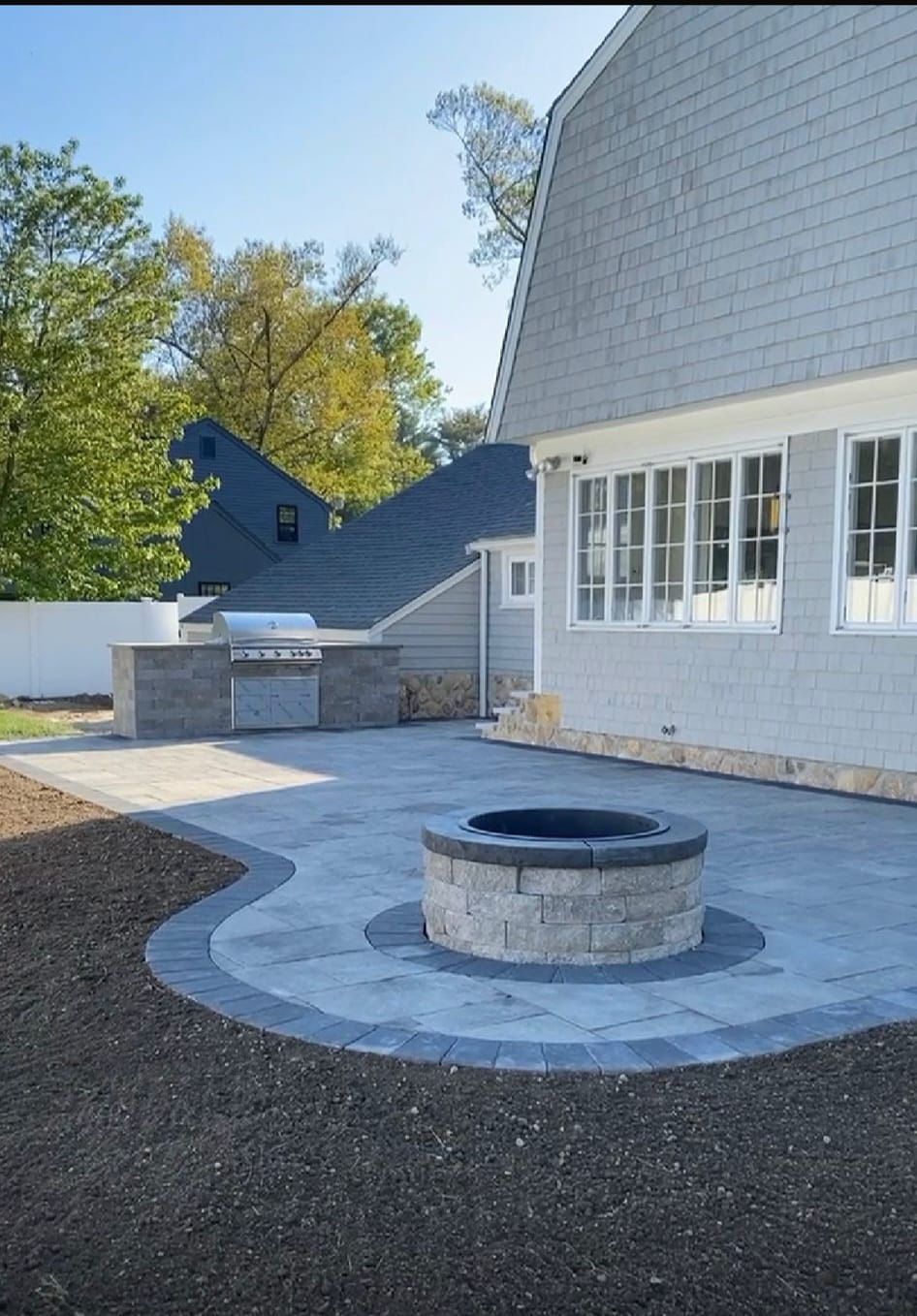 Paver patio with fire pit and outdoor kitchen next to a house with gray siding.