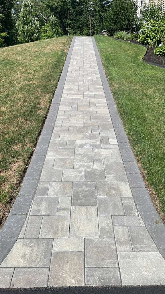 A paved walkway with gray stones bordered by dark gray bricks, leading uphill through a grassy lawn.