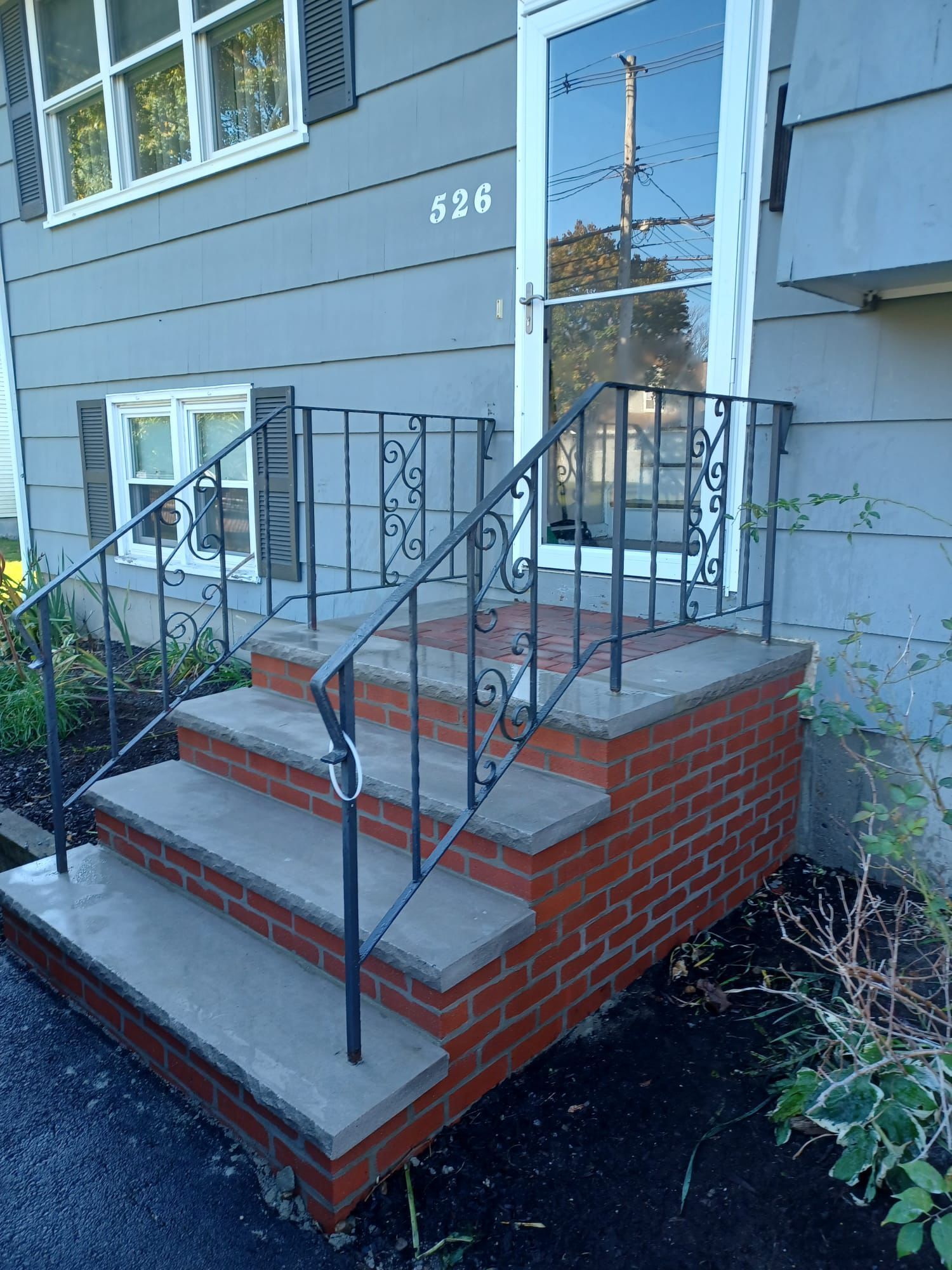 Gray house with three brick steps and ornate black railing leading to a glass door.