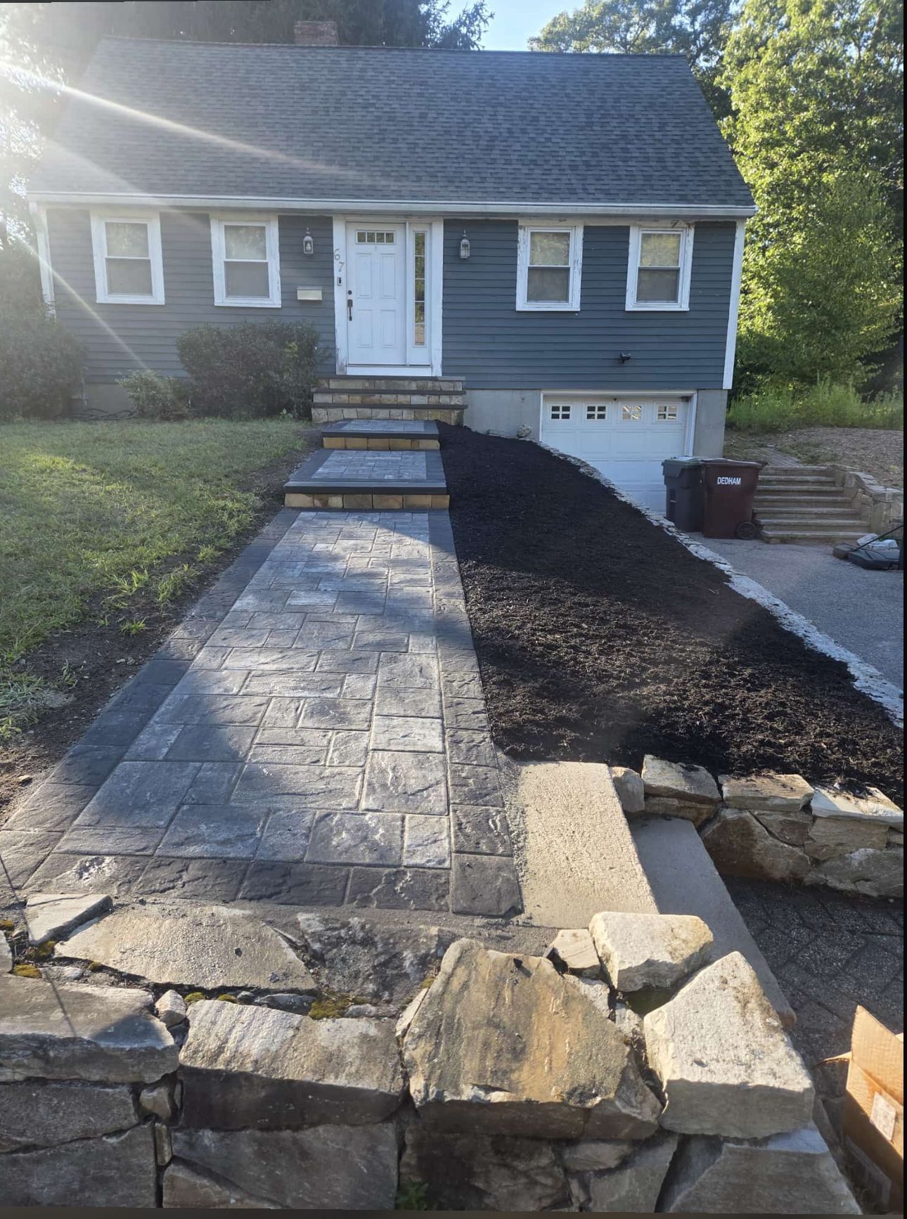 Exterior of a house with a stone pathway, mulch, and rock wall. Gray siding and white door.