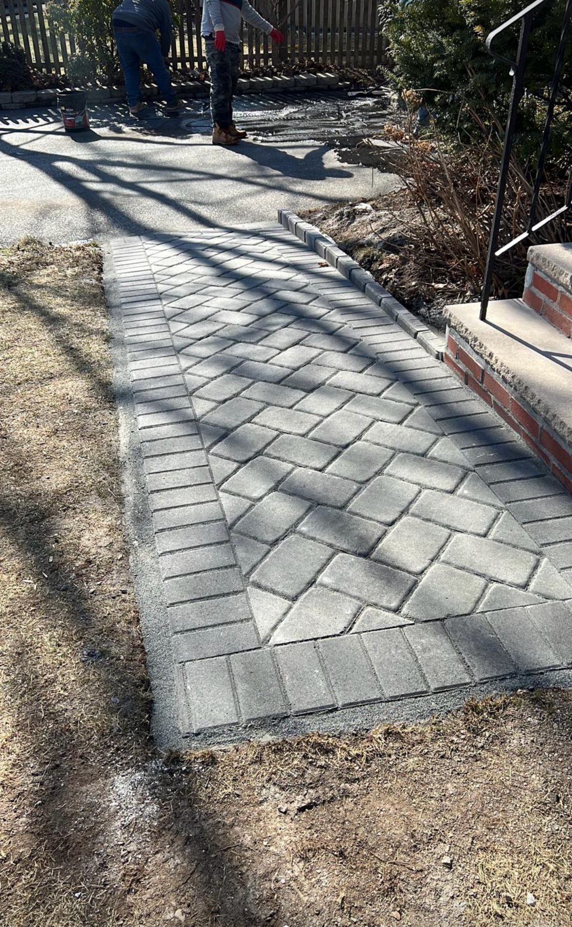 Workers installing a brick paver walkway with a border, leading to a home's steps.