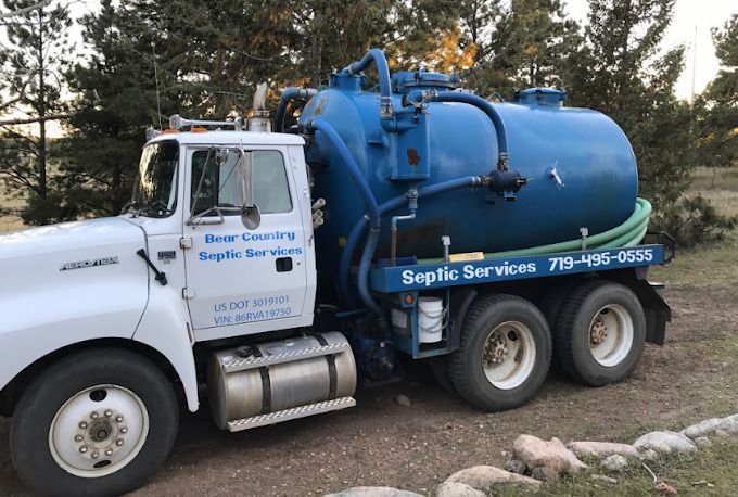 White service truck with blue vacuum tank equipment parked outdoors near trees and rocks