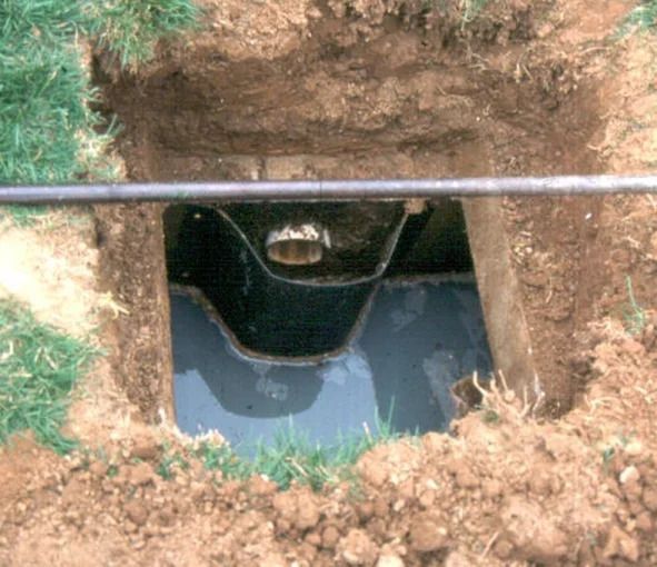 An open excavation revealing a black underground tank with water inside, surrounded by dirt and grass