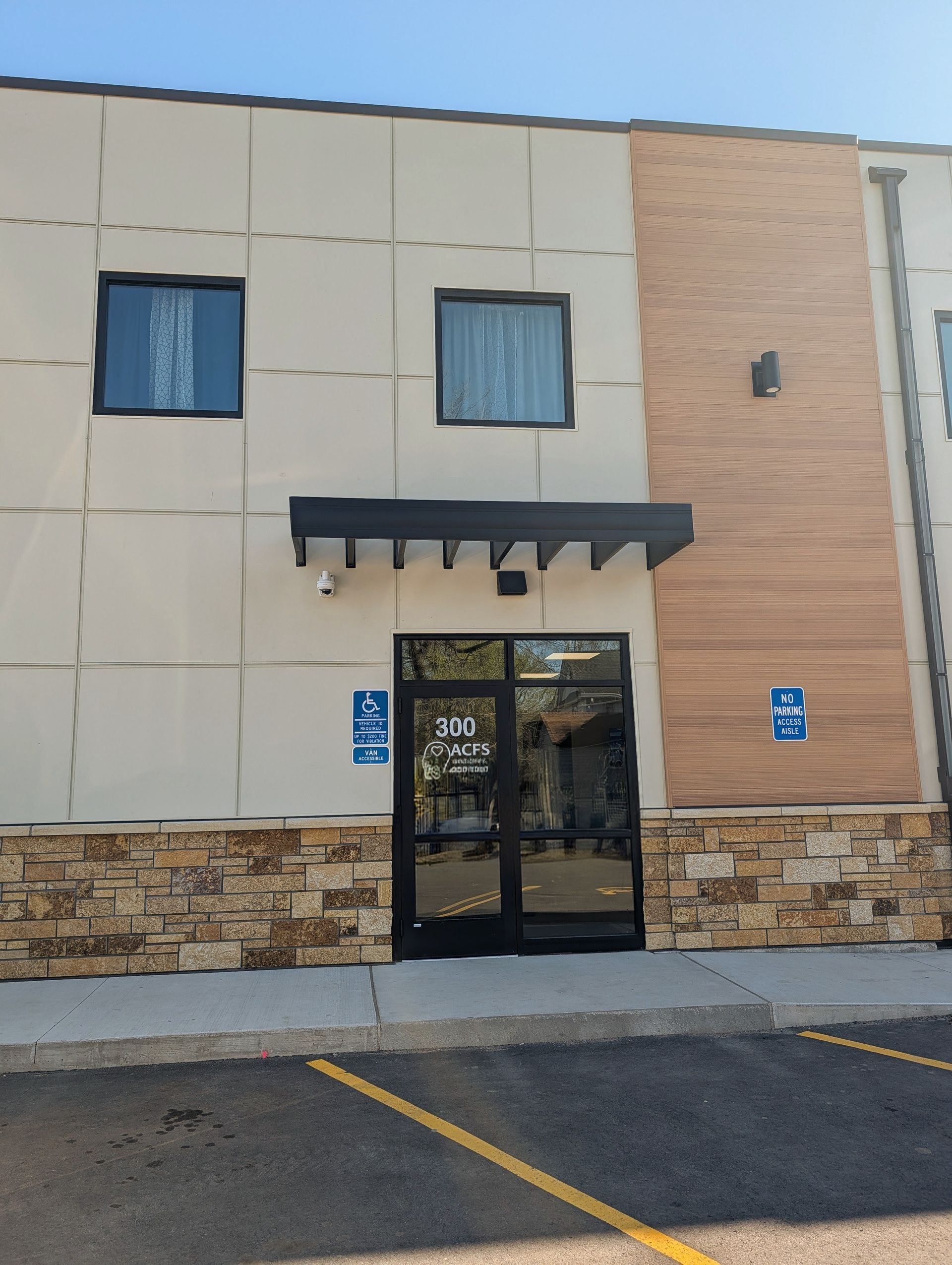 Building entrance with glass doors, beige siding, stone base, and blue accessibility signs by the parking lot