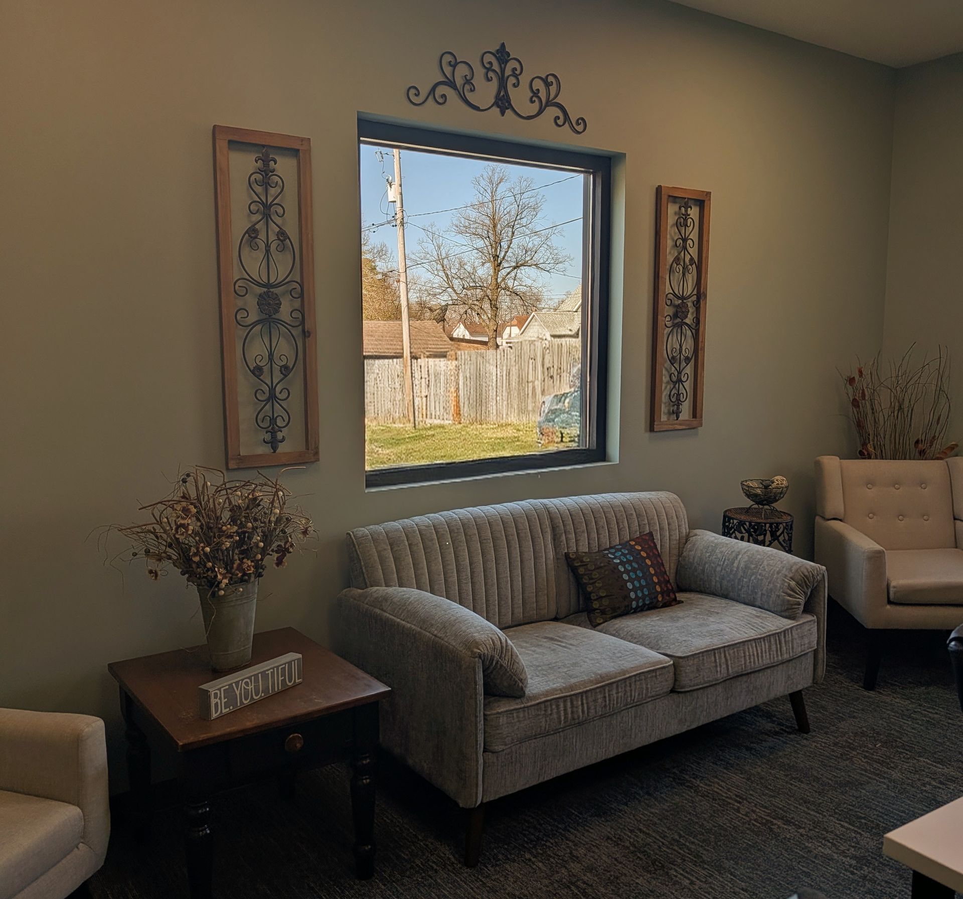Cozy living room with gray sofa, beige armchair, side table, and a window view of a fenced yard.