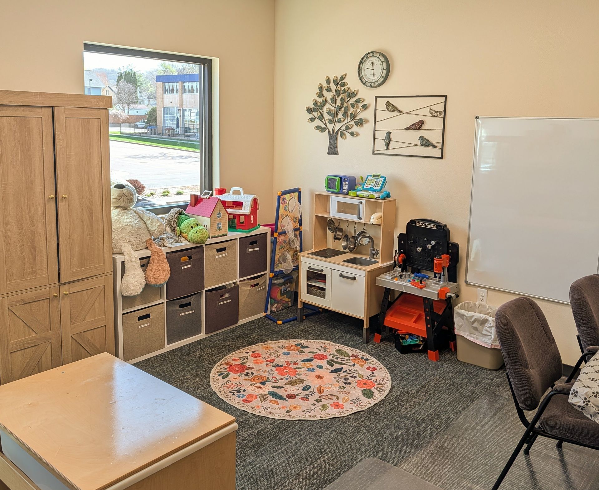 Small classroom corner with cubbies, desk, chairs, whiteboard, rug, window, and wall art.