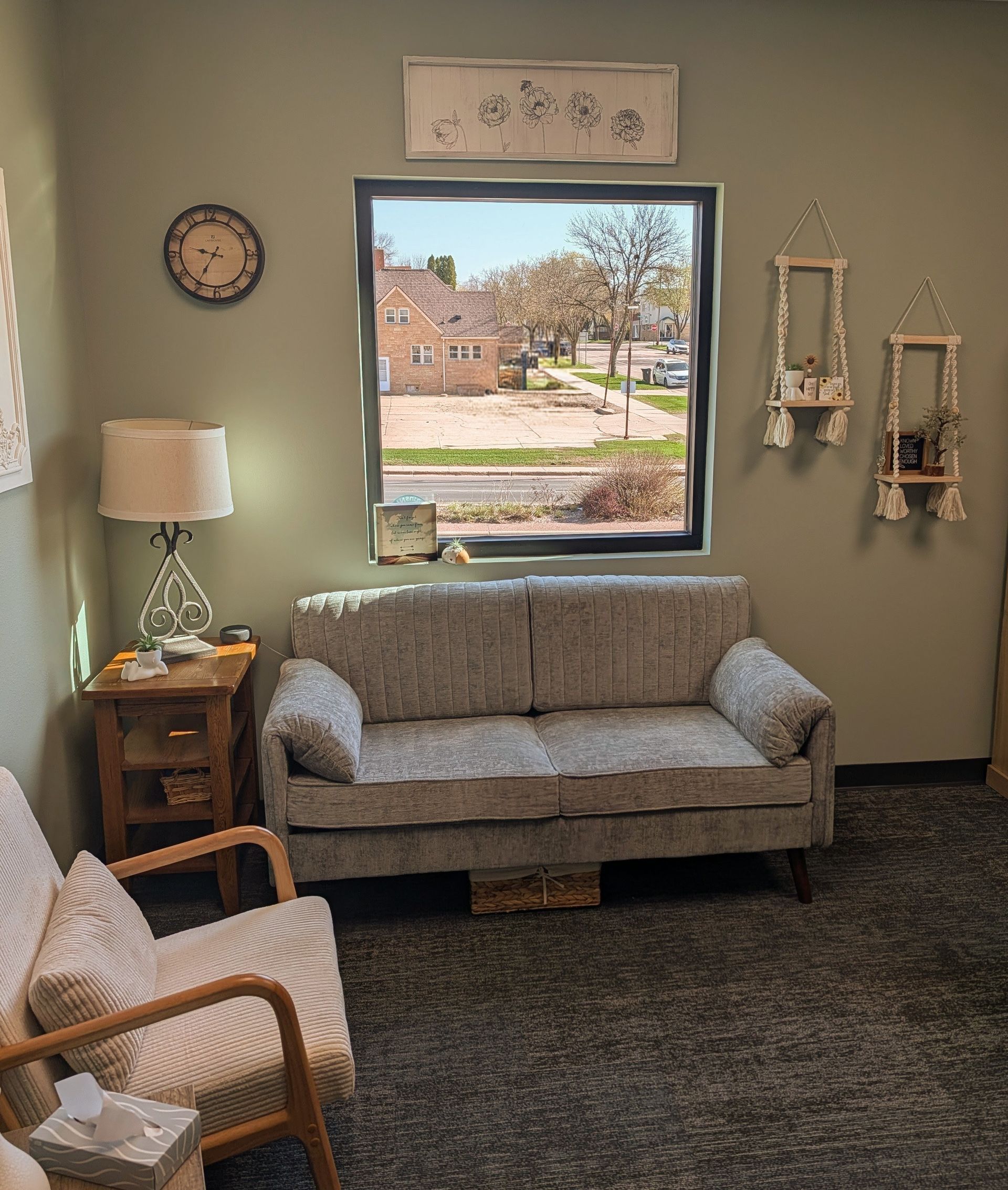 Small waiting room with couch, chair, lamp, clock, and window view of a building and trees.