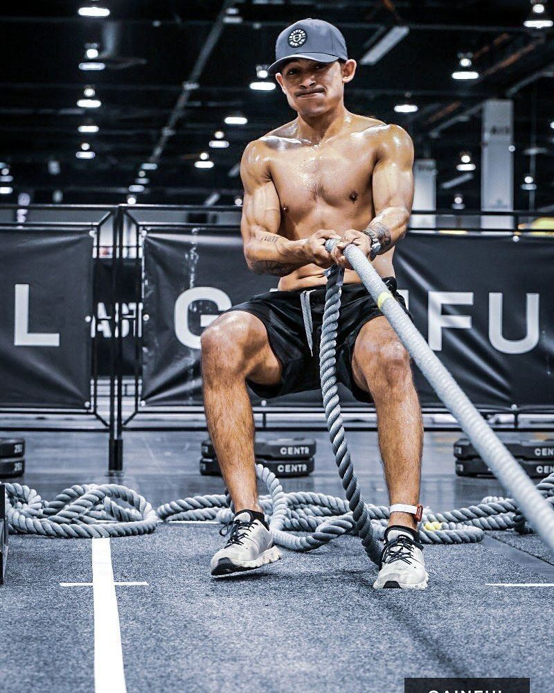A shirtless man is sitting in a gym holding a rope.