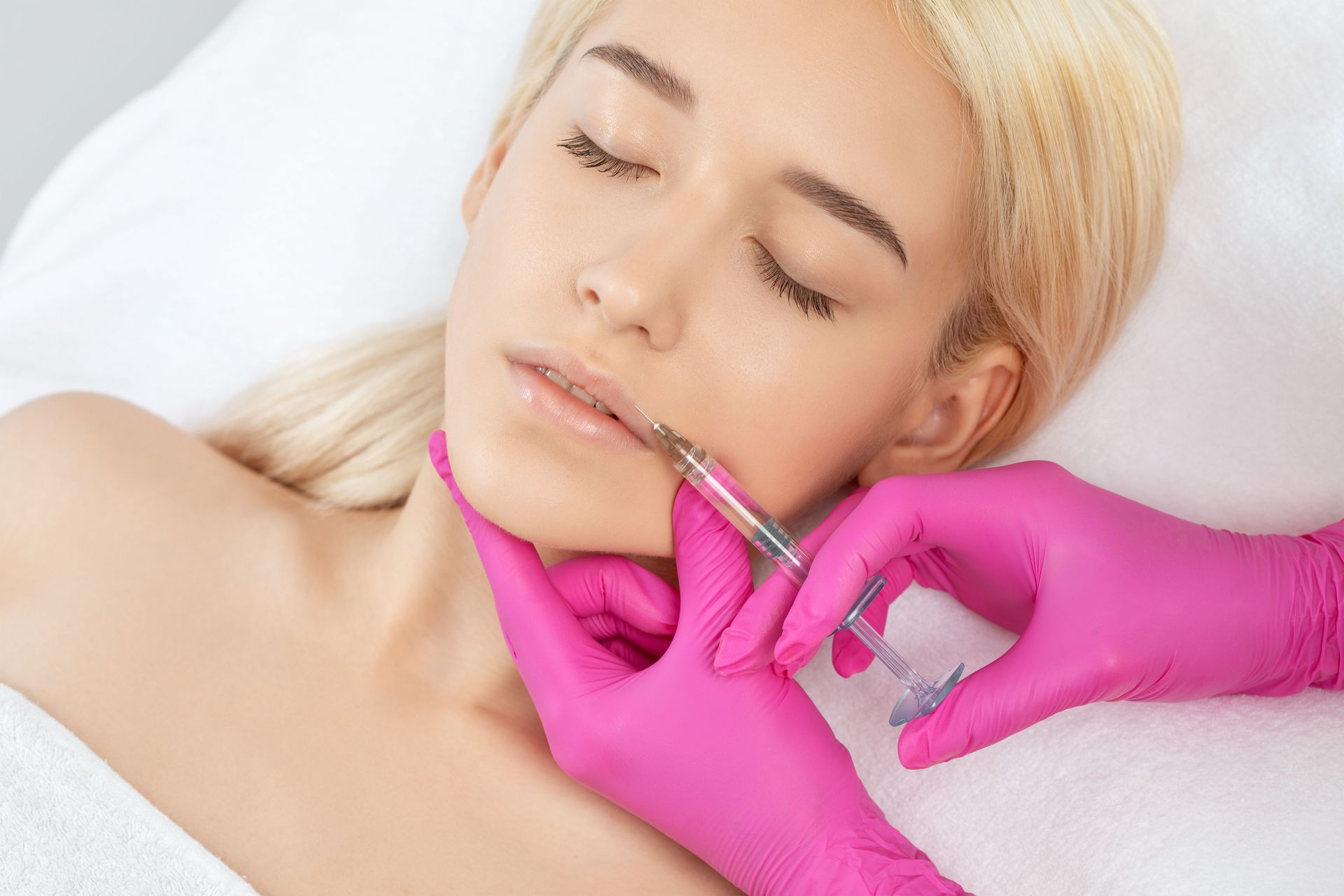 A person in bright pink medical gloves administers a cosmetic lip filler injection to a client lying on a white surface.